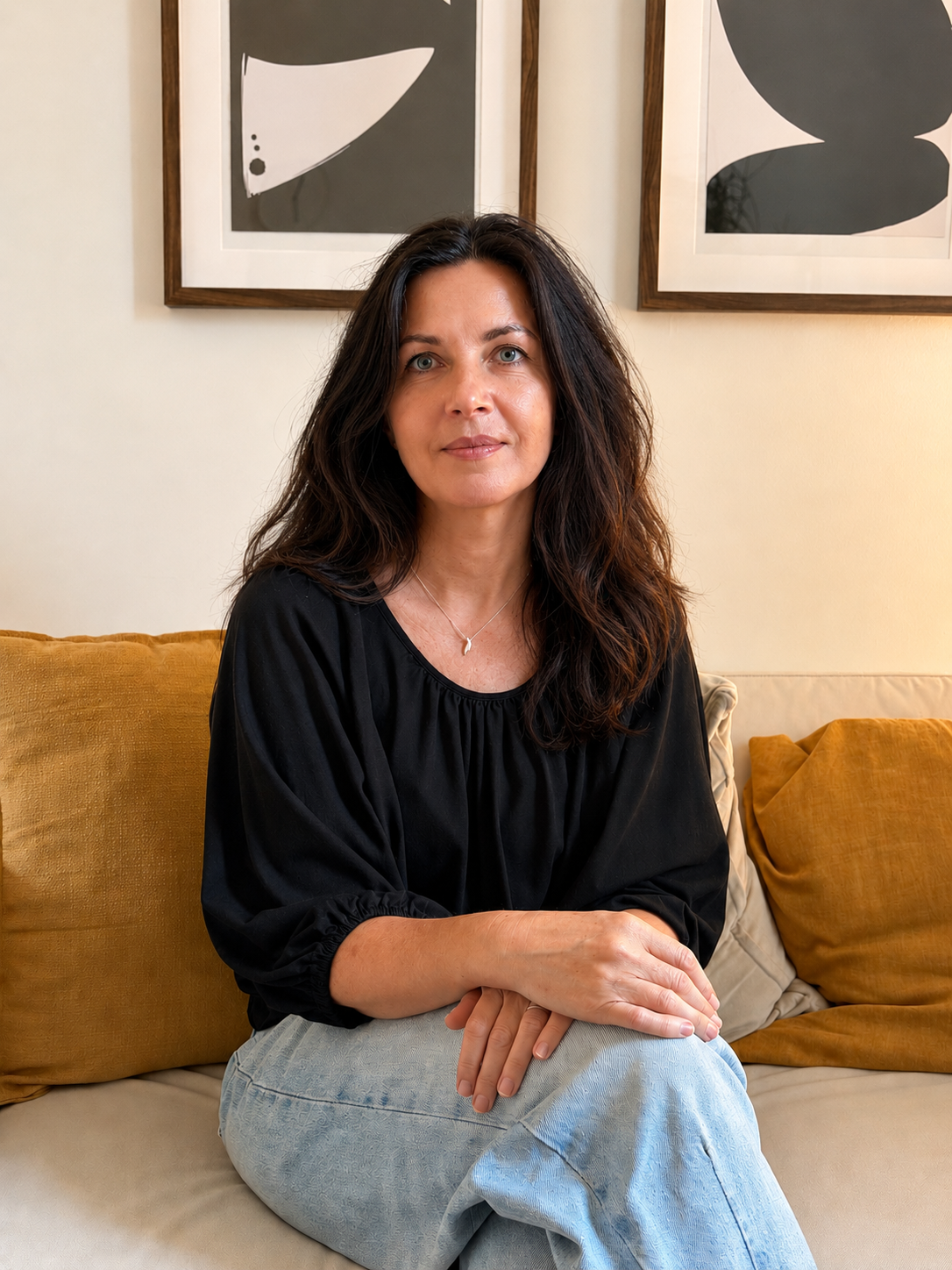 Therapist in Toronto, a woman with dark wavy hair wearing a black top and light blue jeans, sitting on a beige couch with yellow cushions and framed abstract black and white artworks on the wall behind her.