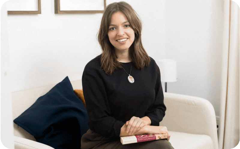 A woman sitting on a white sofa, holding a book, smiling at the camera, in a cozy living room with framed art on the wall. Noema Therapy, List of Therapists, Little Portugal Therapy, Dundas st West therapy