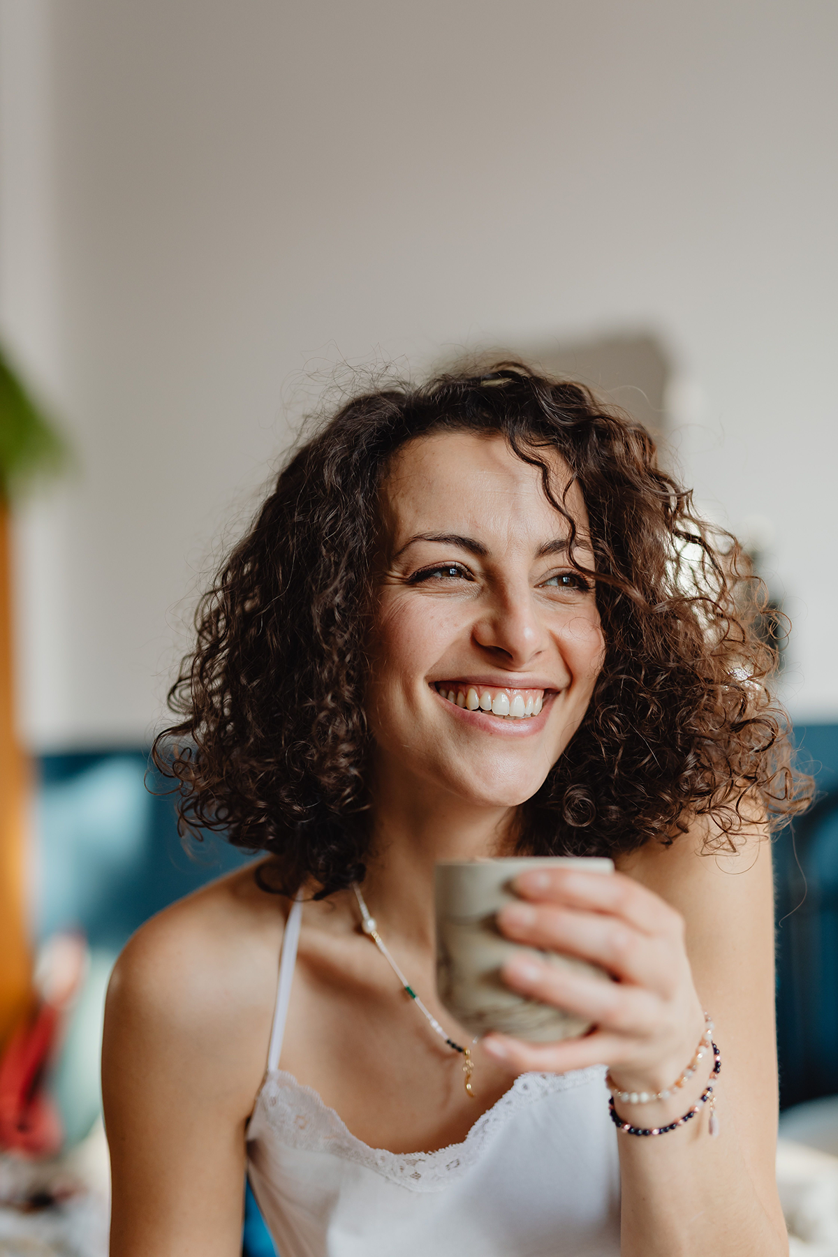 A woman with curly brown hair smiling while holding a beige mug, wearing a white tank top and jewelry.