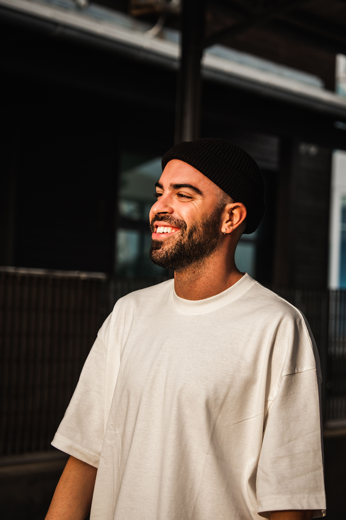 A smiling man with a beard, wearing a black knit cap and a white t-shirt, standing outdoors in an urban setting.