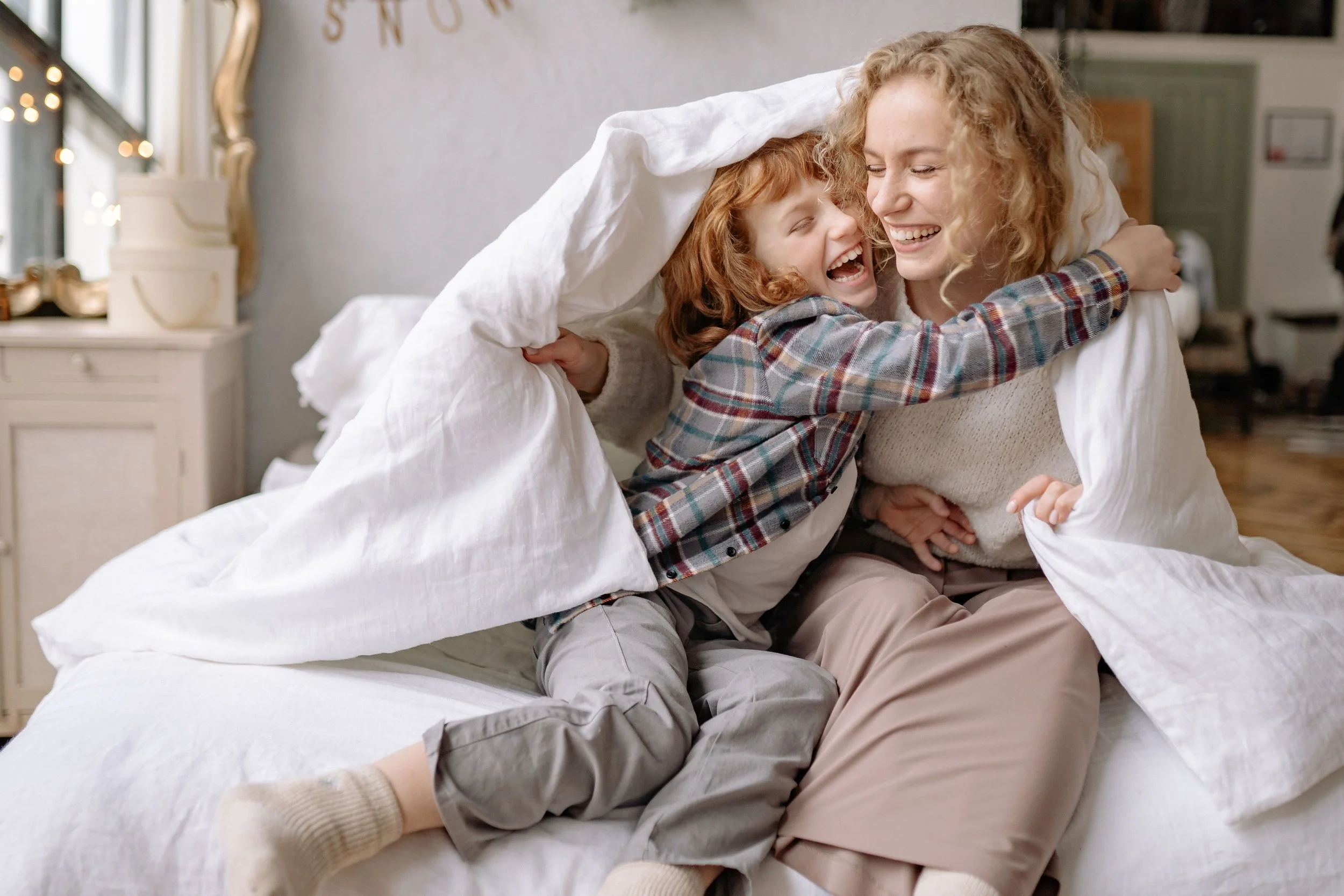 A woman and a young girl sharing a joyful hug on a bed, with the girl sitting on the woman's lap and both smiling, in a cozy bedroom setting.