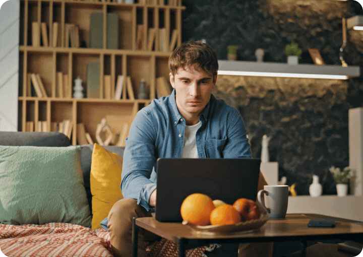 Young man sitting on a couch using a laptop, with a bowl of fruit and a coffee mug on a table in front of him, in a cozy living room with a bookshelf and decorative items. Therapy in Toronto