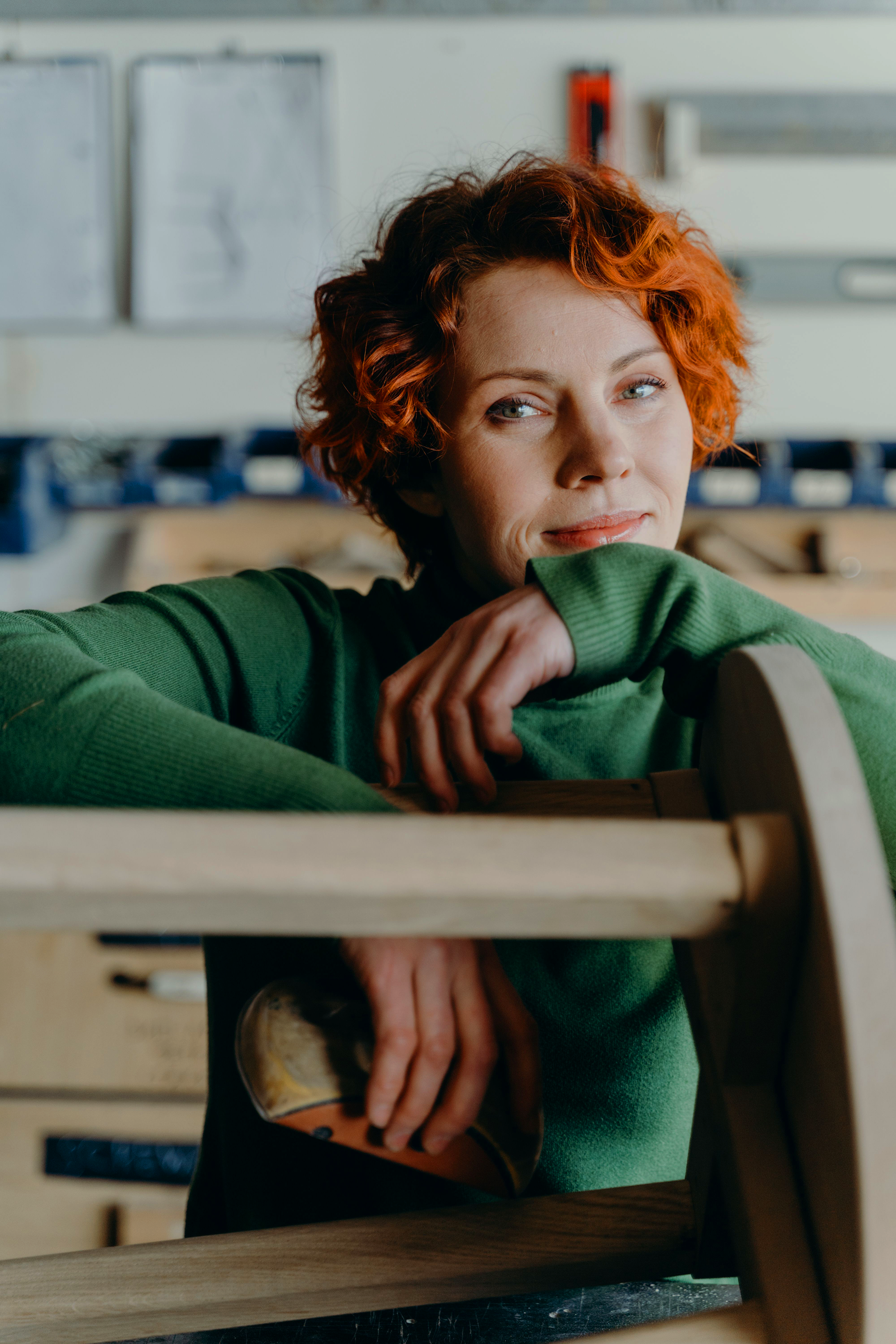 A woman with curly red hair and blue eyes, wearing a green sweater, leaning on a wooden workbench in a workshop or studio.