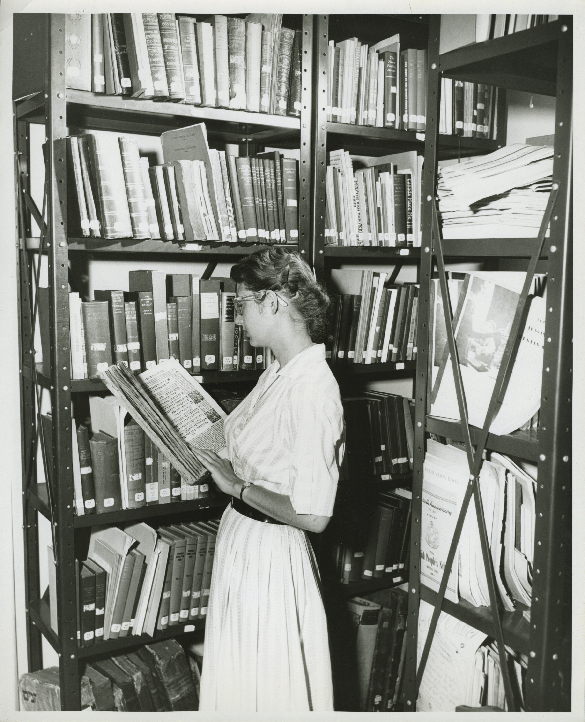 Woman with glasses surrounded by full bookshelves reading an oversized book