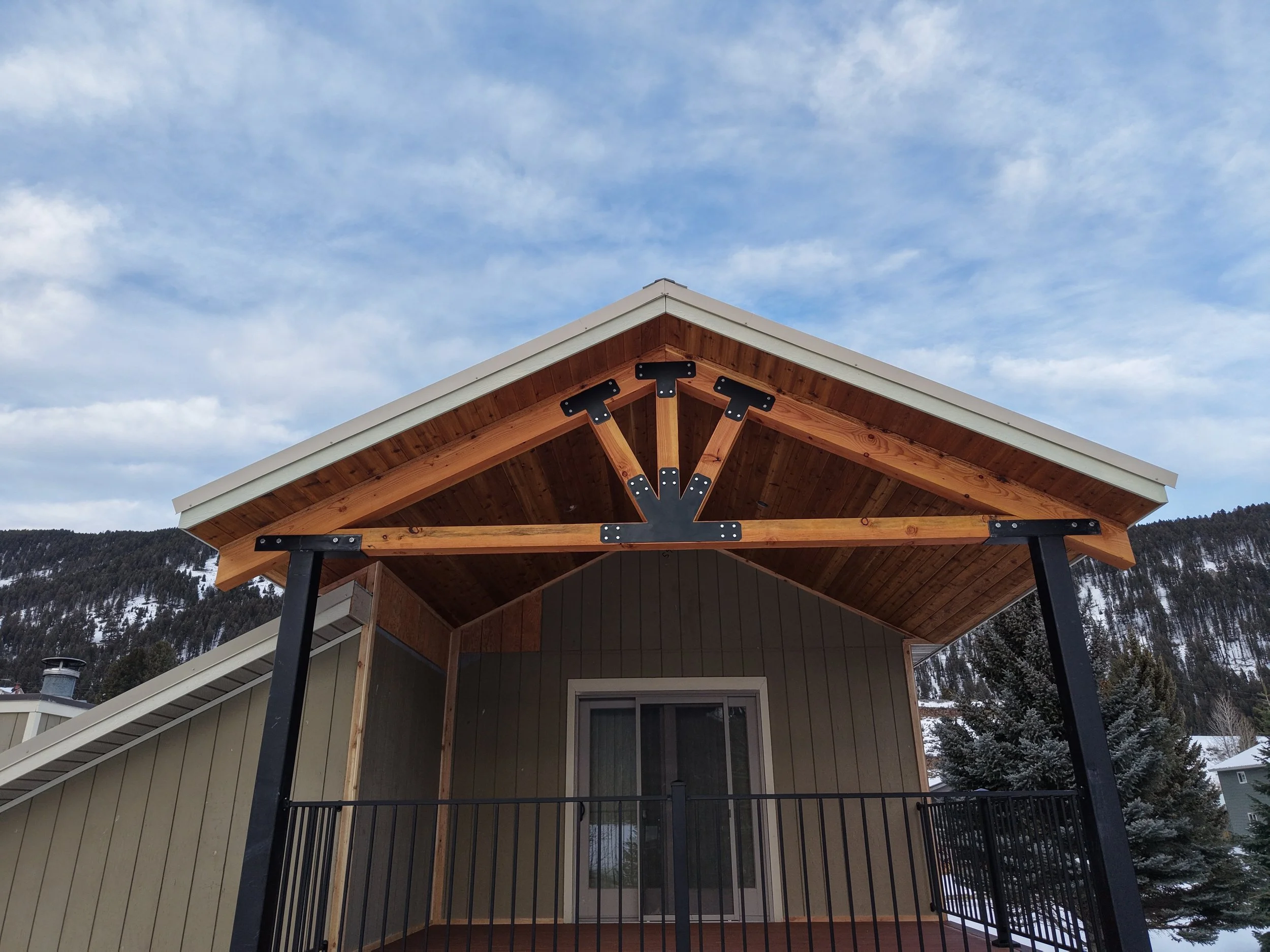 Front view of a house with a wooden porch roof supported by black metal posts. Snow-covered mountains and trees in the background, blue sky with scattered clouds.