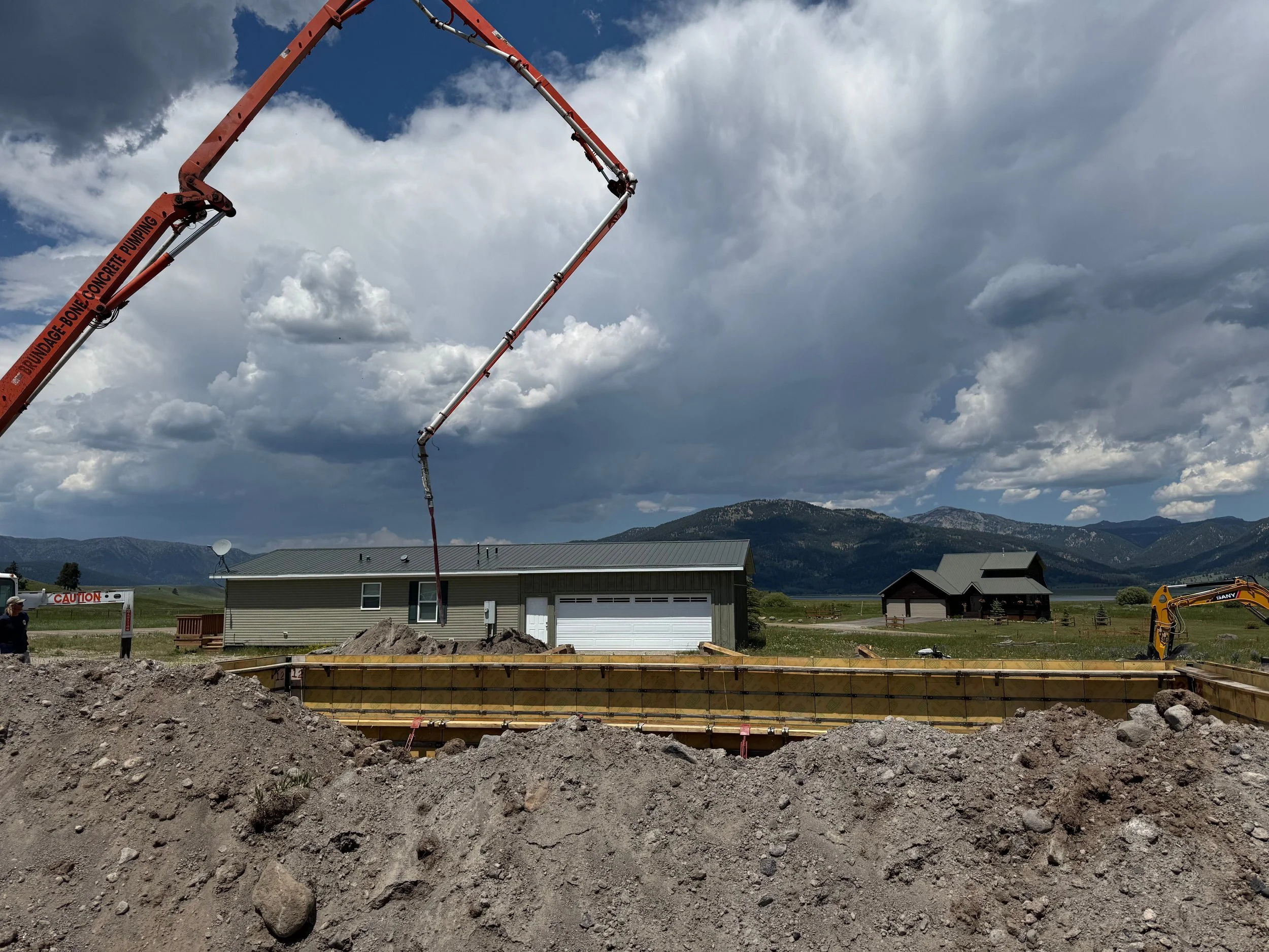 Construction site with a concrete pump, forming a foundation in a rural area with houses and mountains in the background, under cloudy sky.