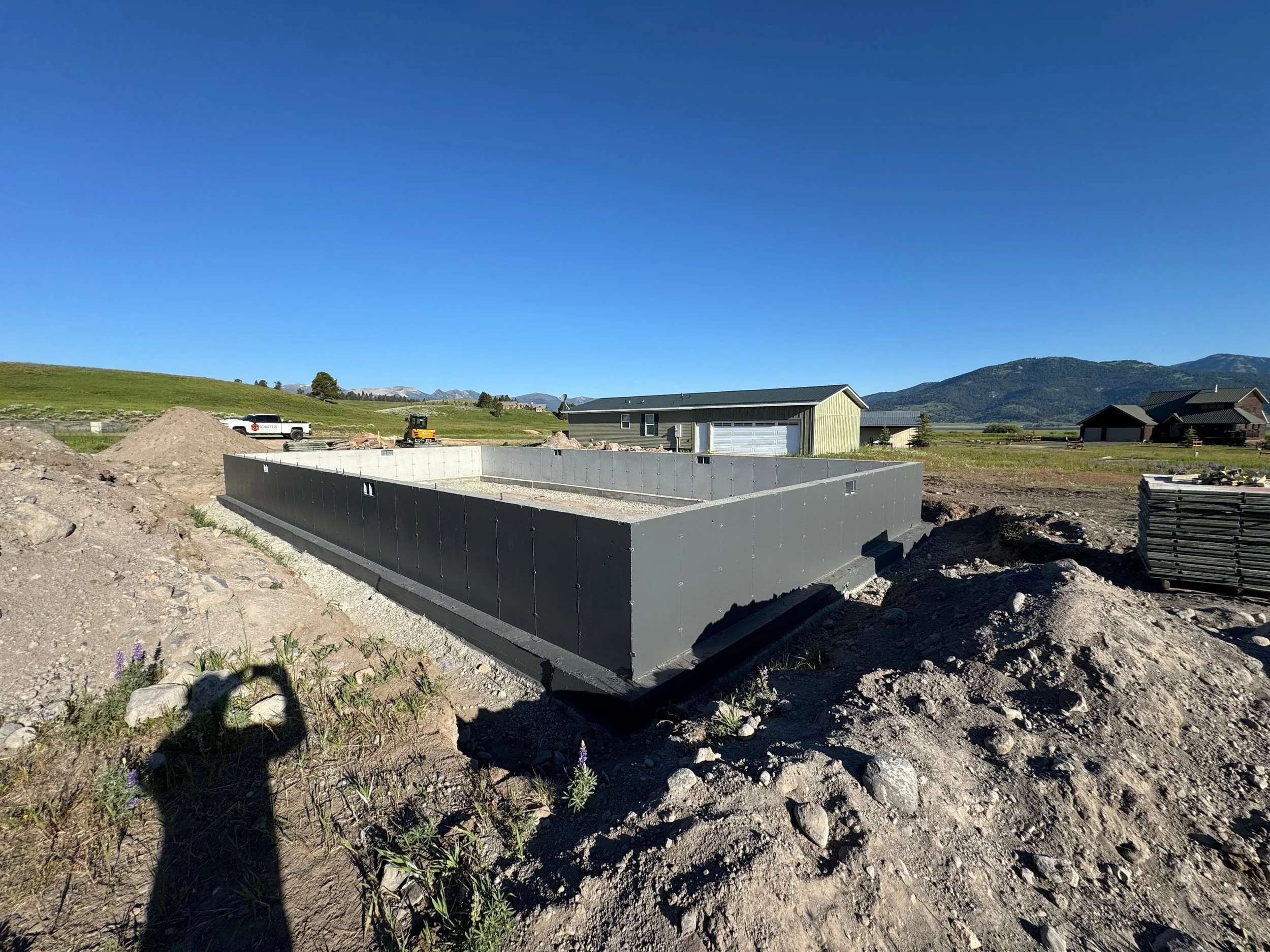 Construction site with a concrete foundation, a small excavator, and a truck, in a rural area with mountains in the background under a clear blue sky.