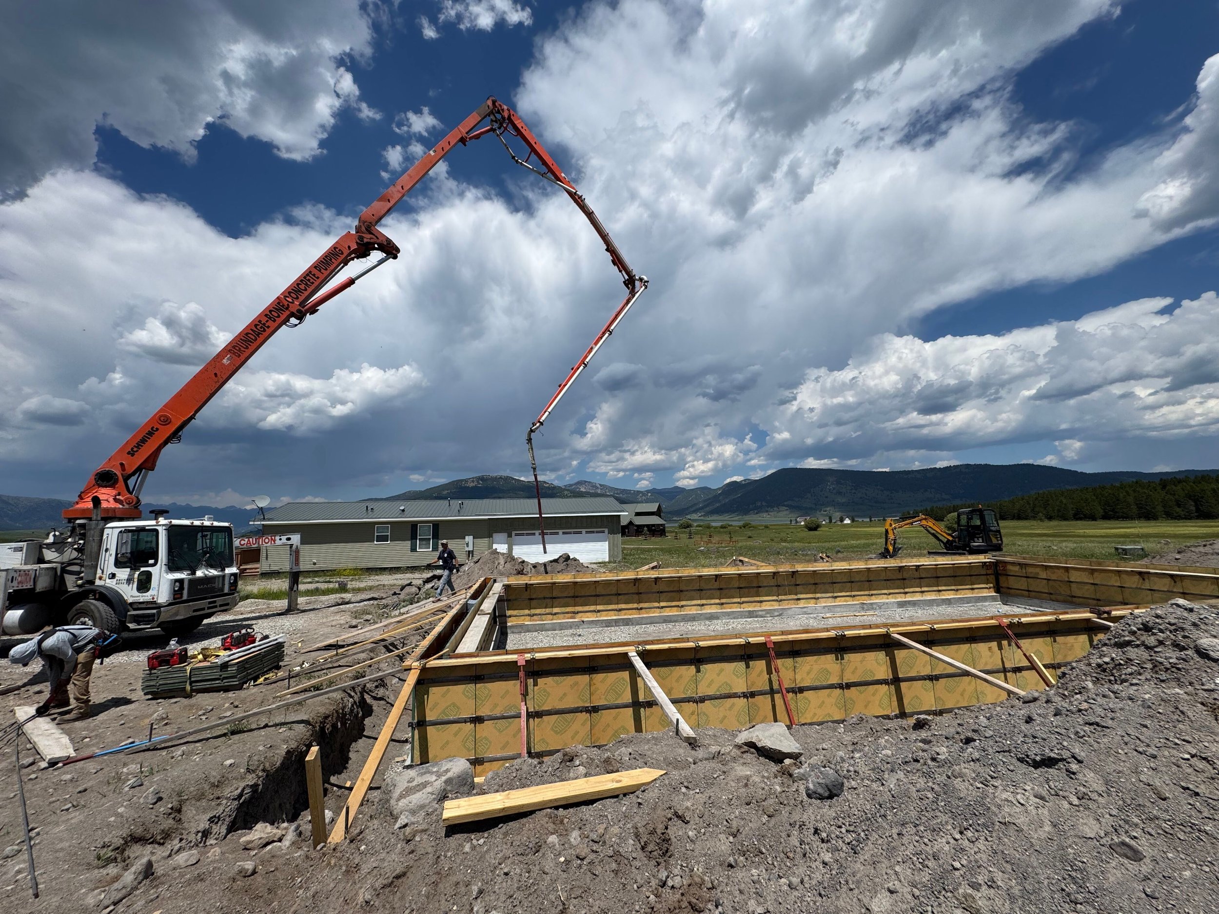 Construction site with a concrete pump truck pouring concrete into a wooden form for a foundation, workers, and smaller machinery against a backdrop of mountains and cloudy sky.