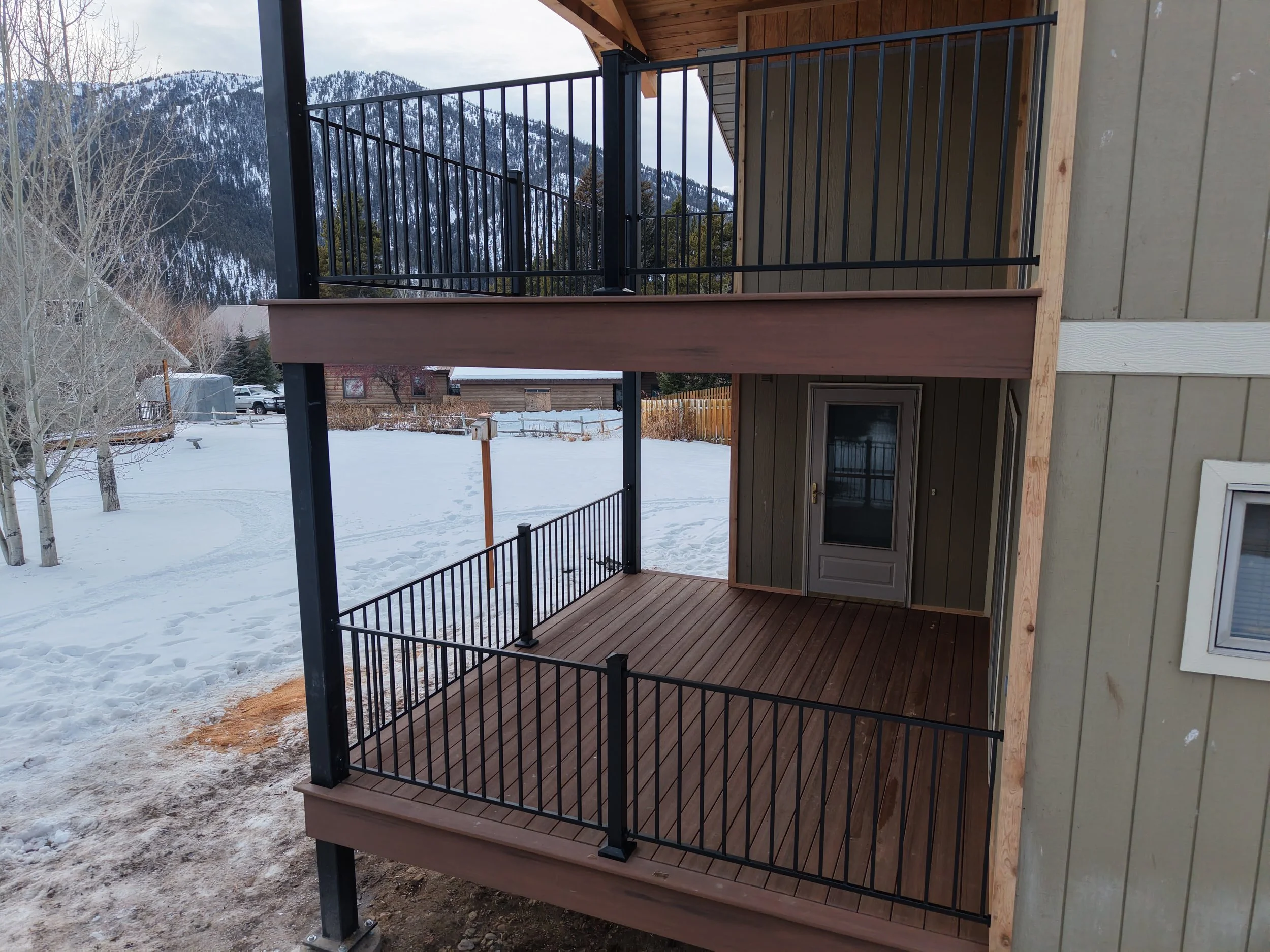 New wooden balcony with black railings attached to the exterior of a house, overlooking a snowy yard with mountains in the background.
