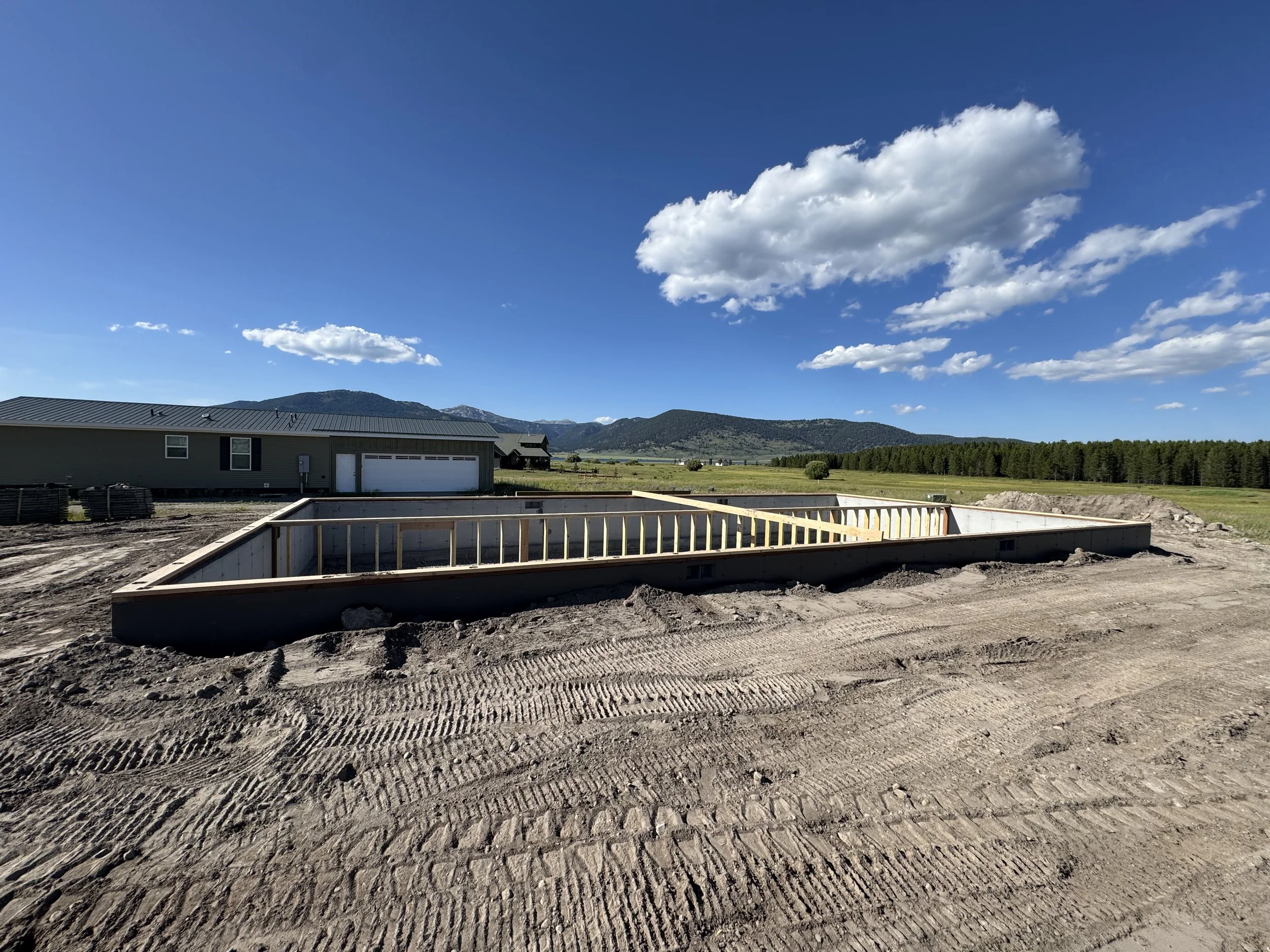 Construction site with a partially built house foundation, surrounded by dirt and tire tracks, with green fields, trees, and mountains in the background under a blue sky with scattered clouds.