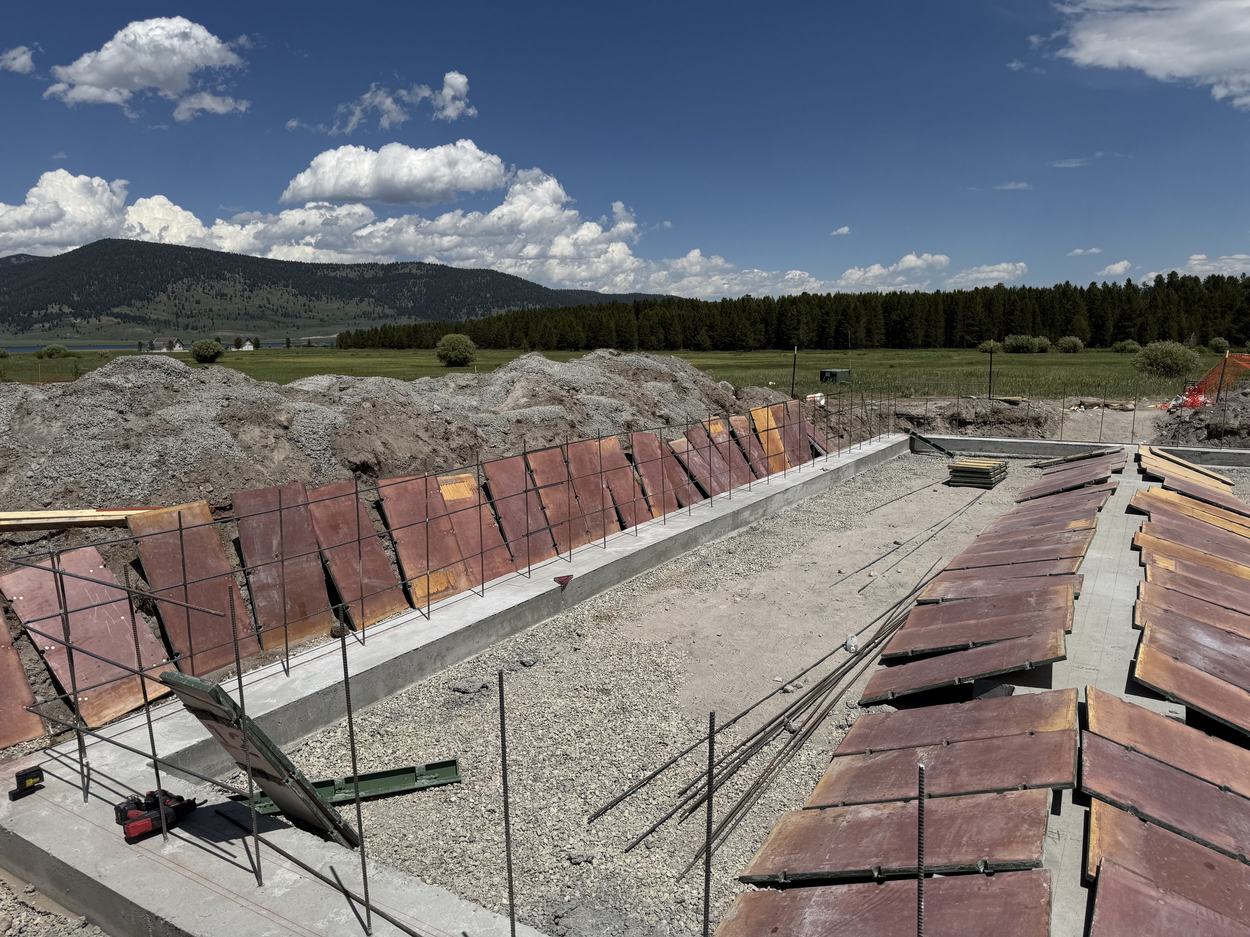 Construction site with a concrete foundation and red roofing tiles, surrounded by dirt piles, in a rural area with green fields, trees, and mountains in the background under a partly cloudy sky.