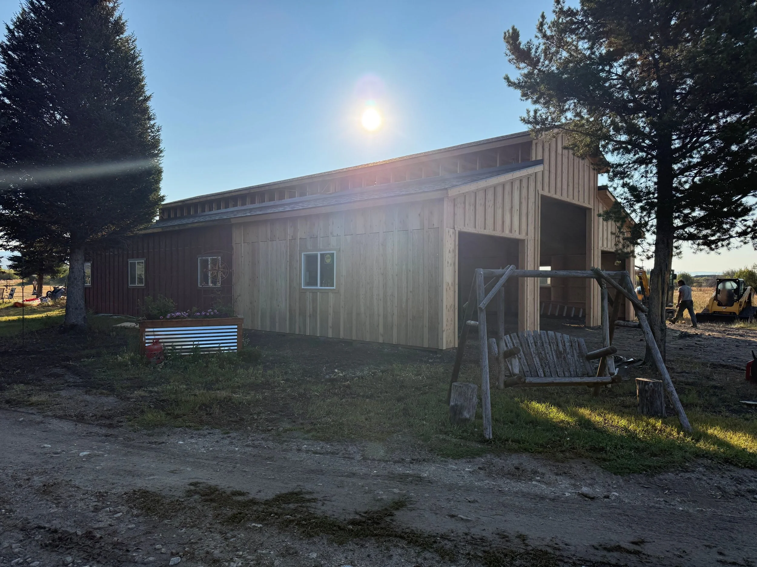 A wooden building under construction with a metal roof, surrounded by trees, a swing, and construction workers in the background, with the sun shining overhead.