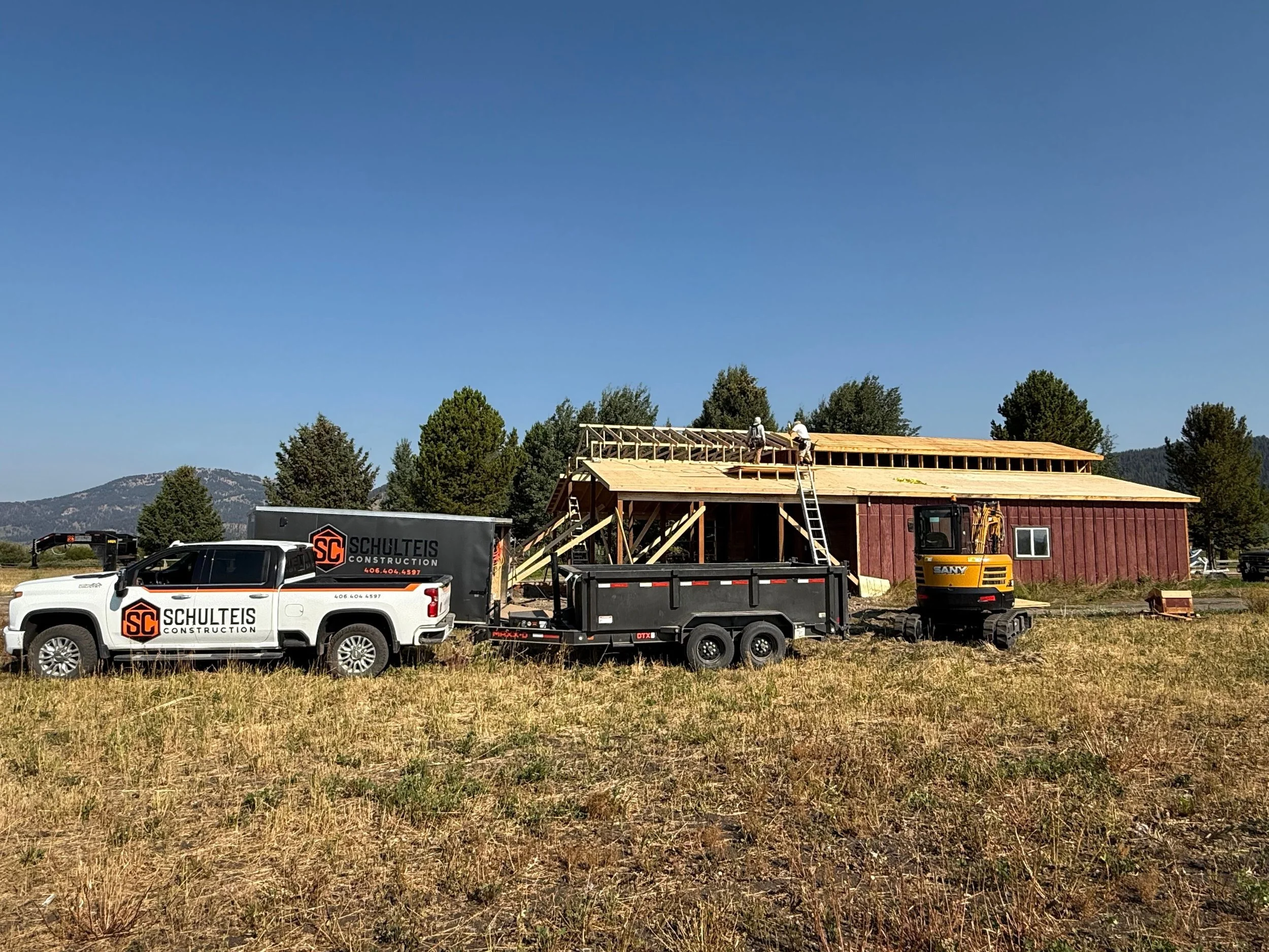 Construction site with a building under construction, a white pickup truck with 'Schulteis Construction' logo, a yellow excavator, and workers on the roof. The setting is a grassy field with trees and mountains in the background under a clear blue sk