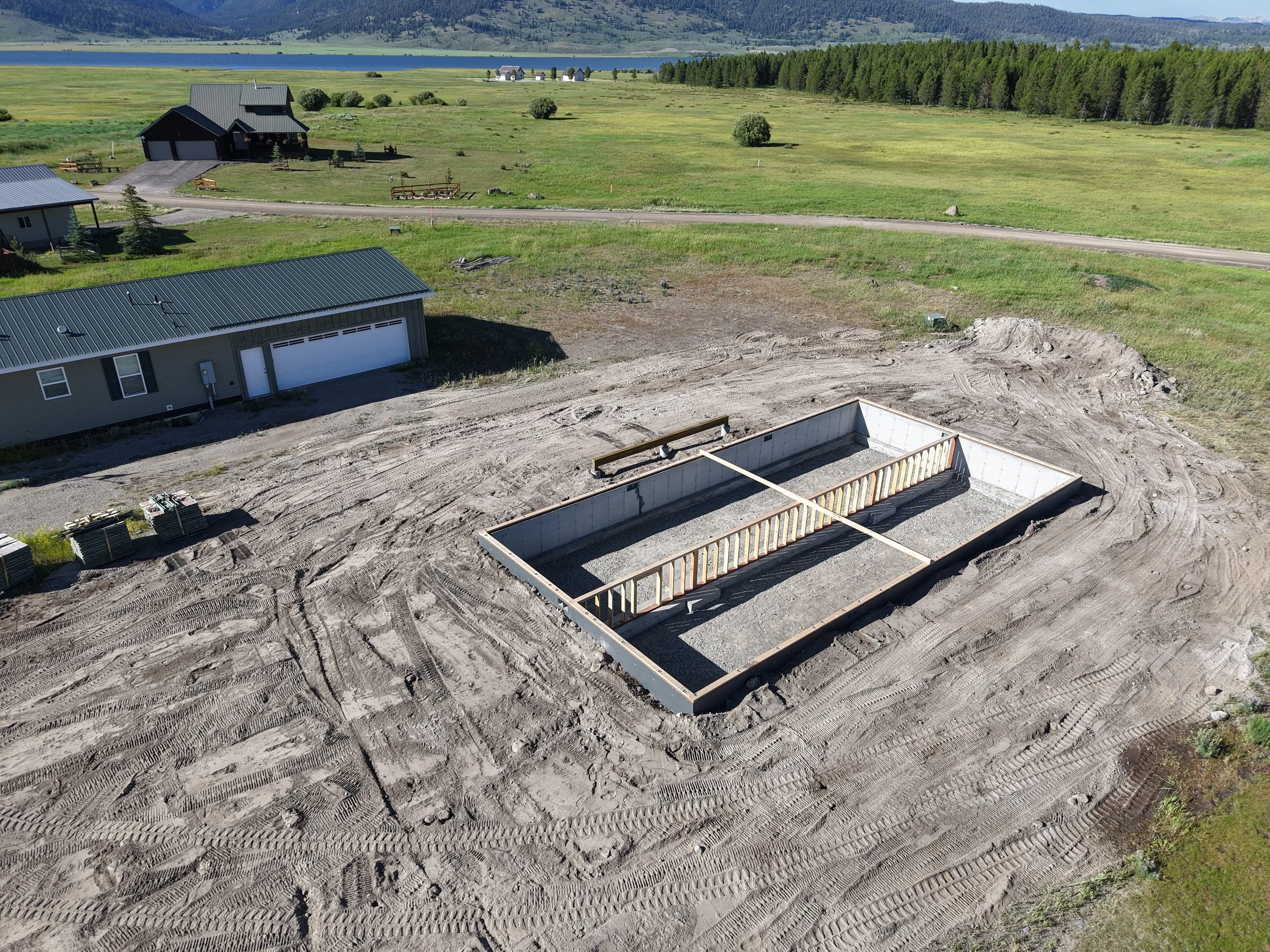 A construction site with a foundation for a new building, surrounded by dirt and tire marks, with a nearby house and green fields in the background.