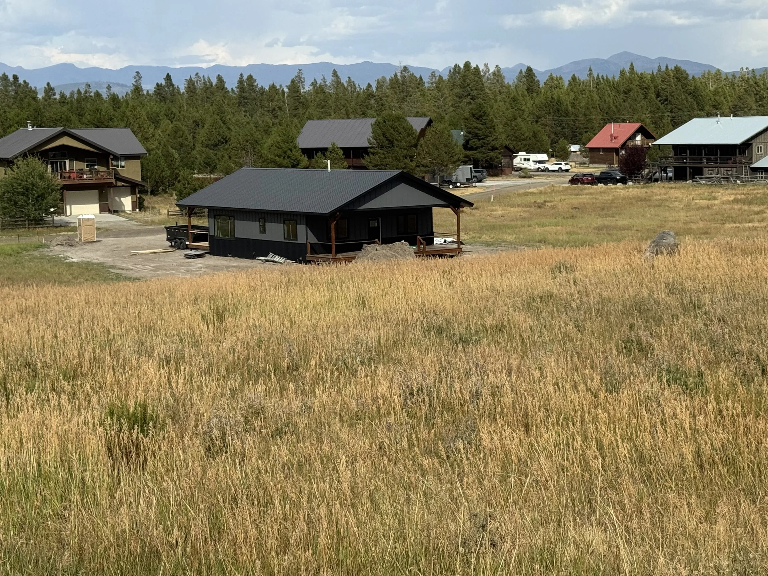 A black house with a porch and gray roof situated in a field of tall grass, with a few rocks visible, and other houses with trees and mountains in the background.