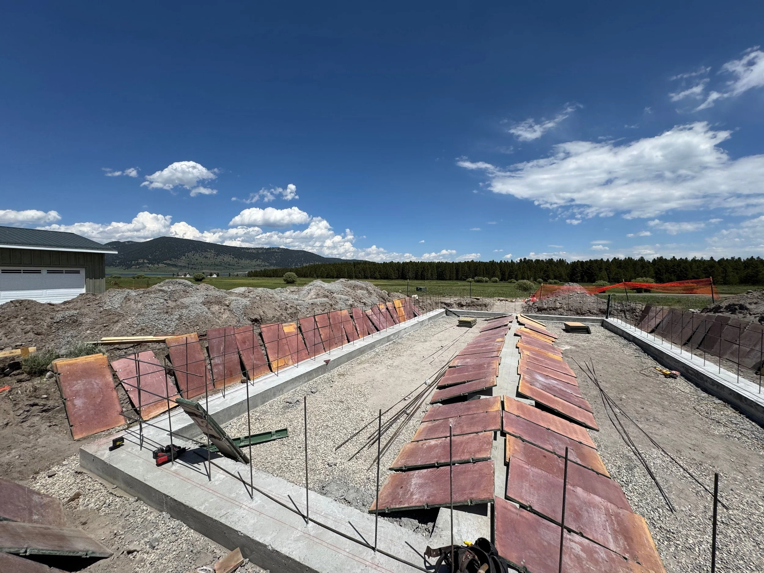 Construction site with a partially built structure featuring red tiles, dirt piles, and a mountainous landscape under a blue sky with clouds.