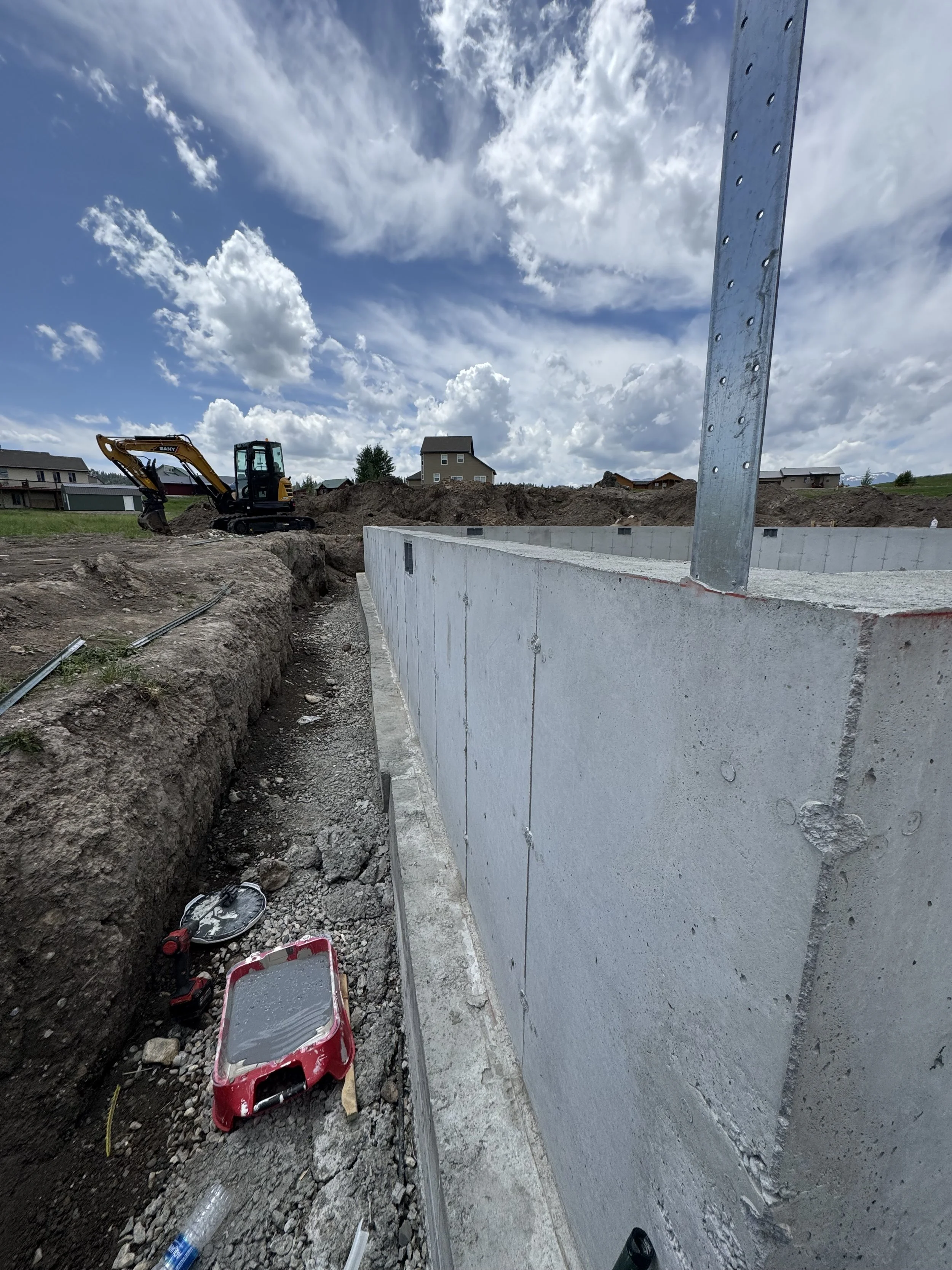 Construction site with a concrete foundation wall and a small excavator working on dirt; partly cloudy sky and residential houses in the background.