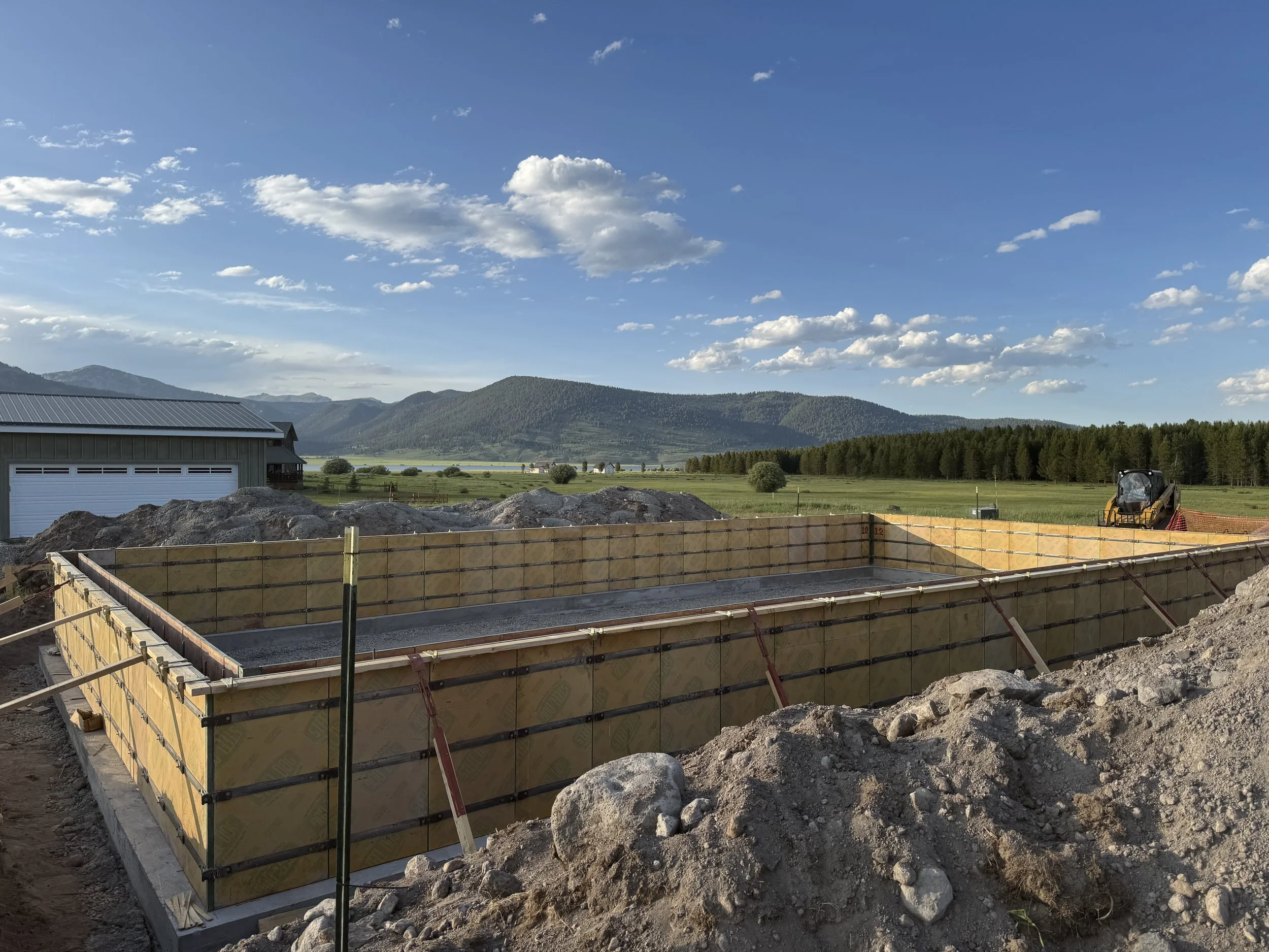 Construction site for an in-ground pool with wooden formwork, piles of dirt and rocks in the foreground, a barn, open fields, trees, mountains, and a partly cloudy sky in the background.