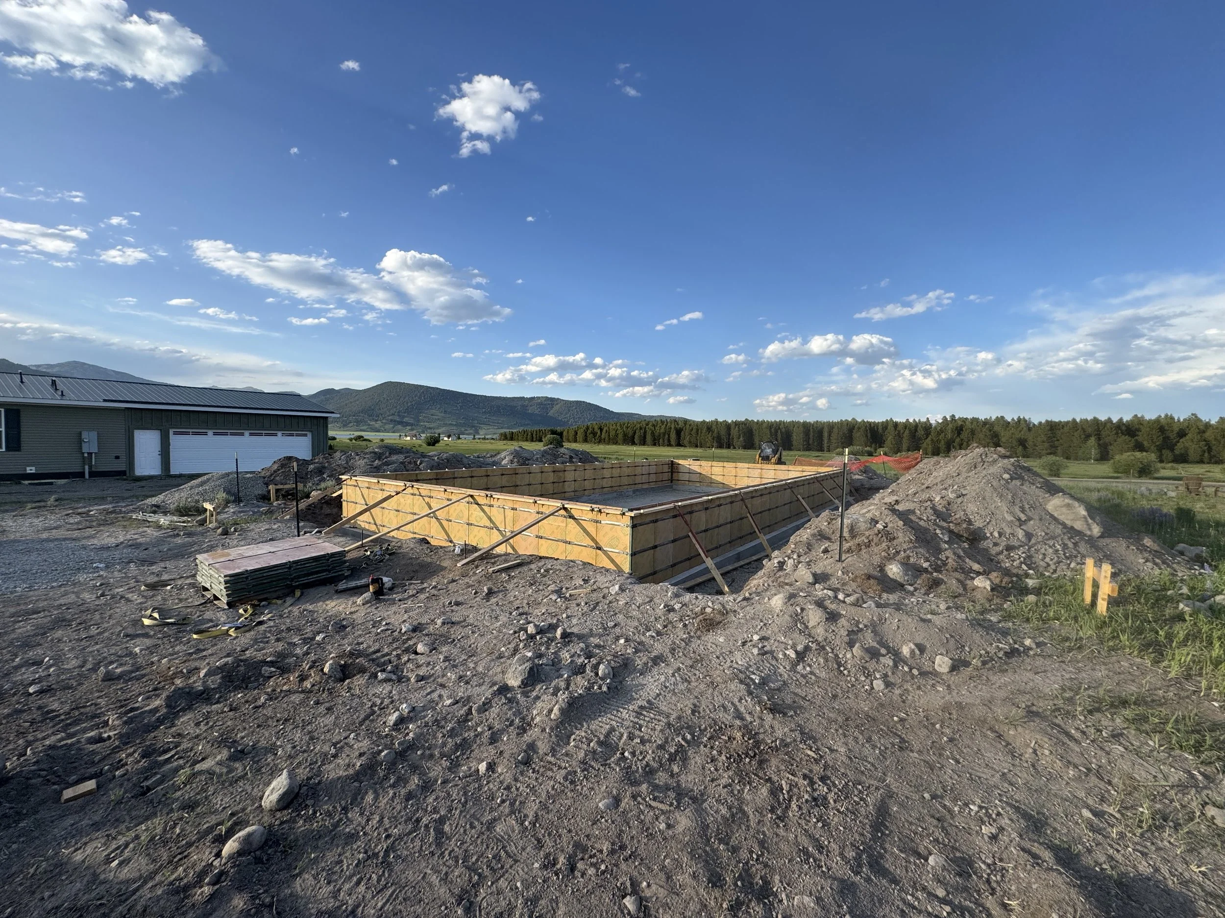 Construction site with a foundation being built, surrounded by dirt and gravel, with a house and mountains in the background under a partly cloudy sky.