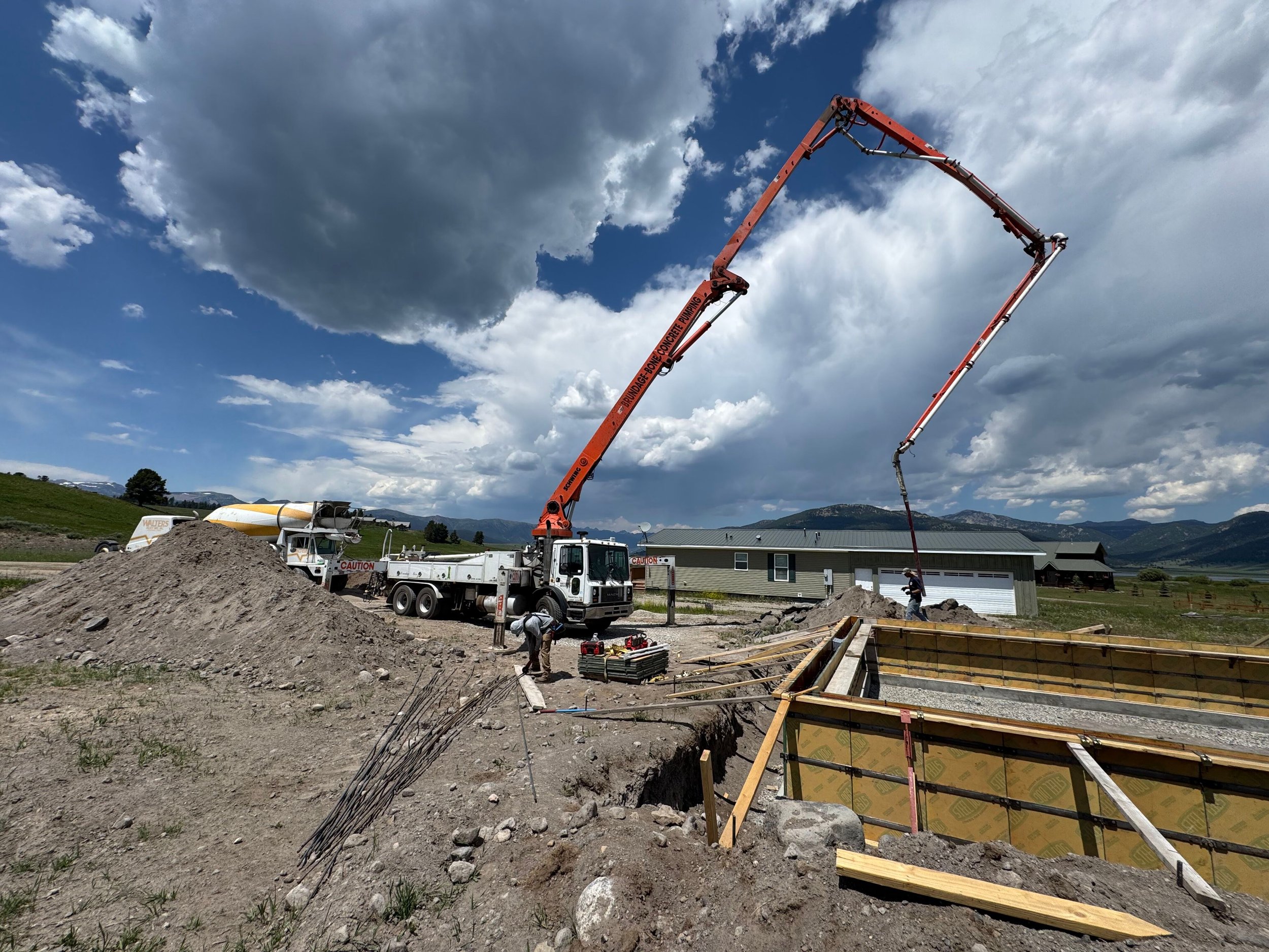 Construction site with a concrete pump truck pouring concrete into a foundation, workers and building materials nearby, rural landscape with hills and clouds in the sky.