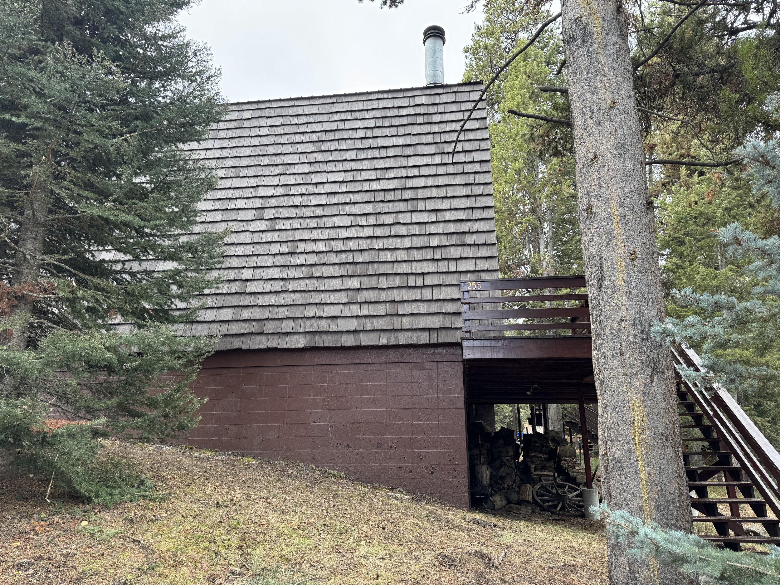 A rustic wooden house with a sloped shingled roof and a raised deck on the right side, surrounded by tall trees in a forested area.