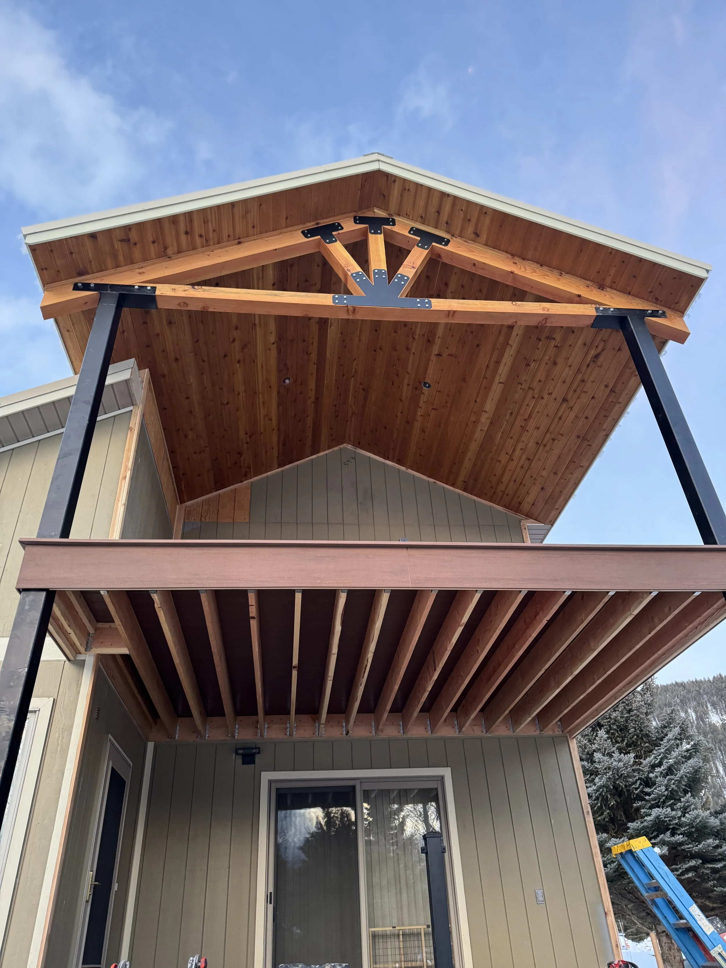 Construction of a multi-level house deck with a upper covered balcony with a wooden ceiling and metal support beams. A blue ladder is visible on the right side.