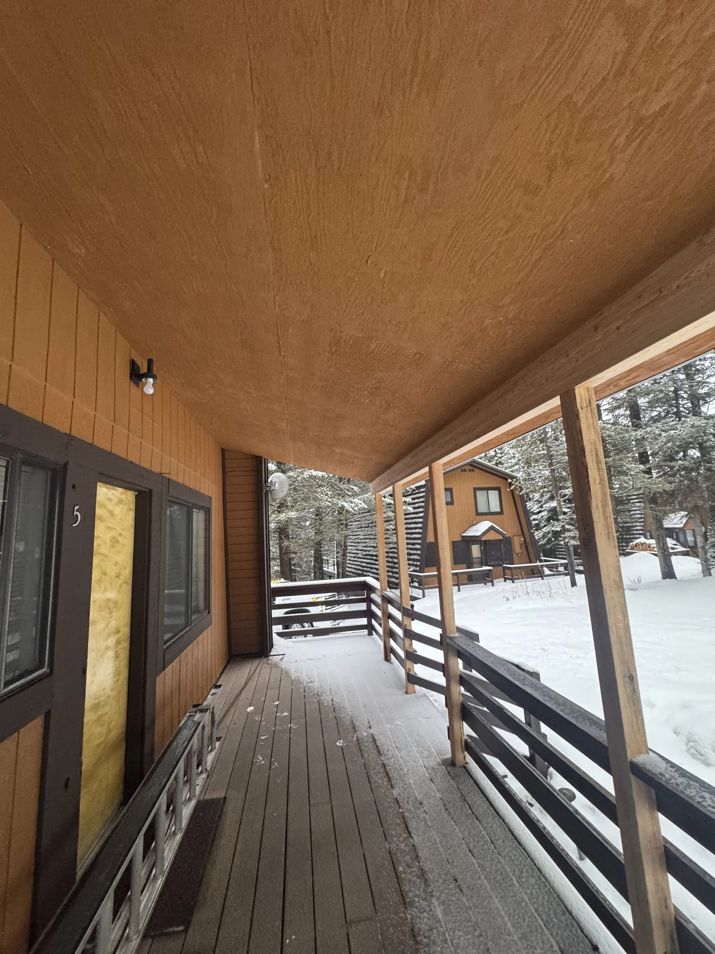 Front porch of a wooden house with snow on the ground, a ladder leaning against the wall, and neighboring houses and snowy trees visible in the background.