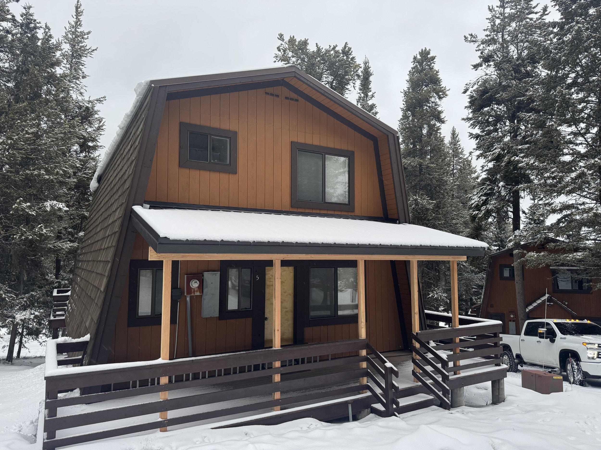 A modern two-story wooden house with a snow-covered roof, in a snowy forested area, with a truck parked in front.