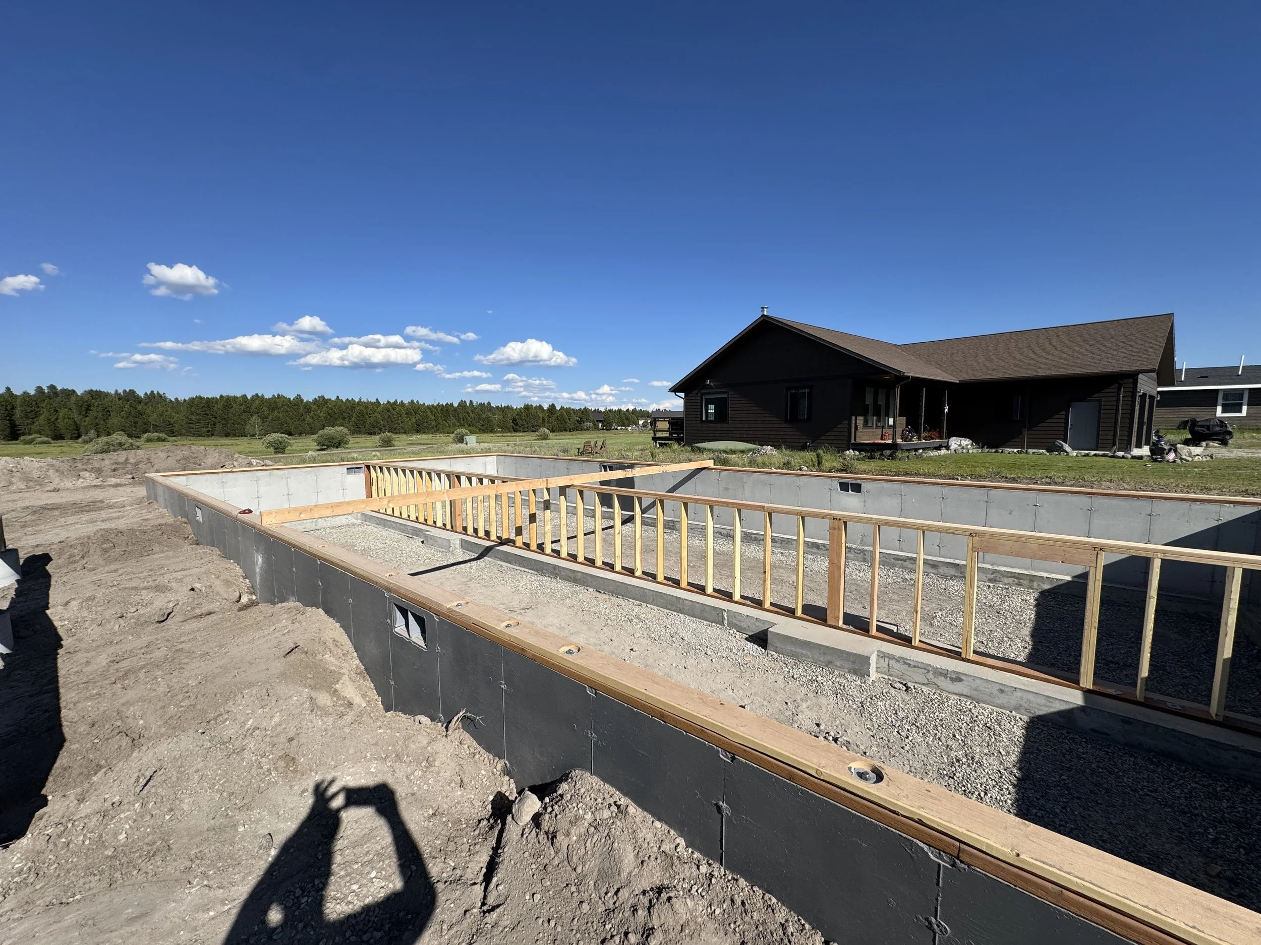 Construction site with building foundation and wooden framing, house in background, clear blue sky, and shadow of person taking photo.