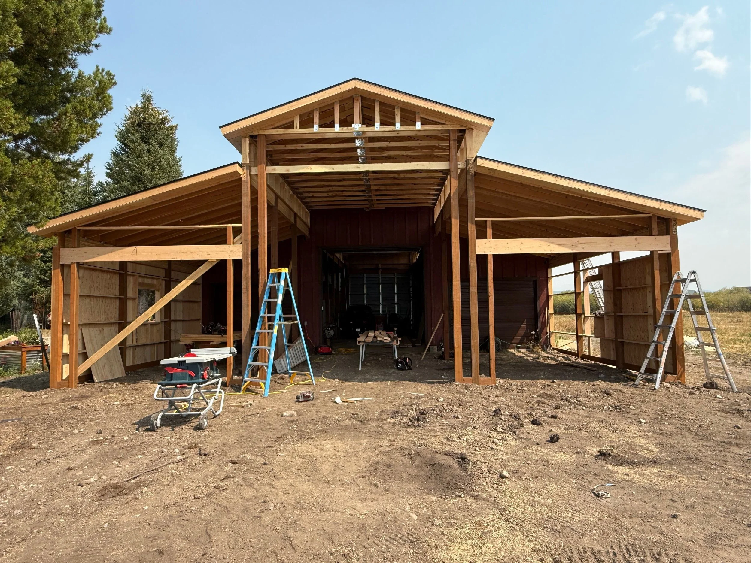 Under construction barn with wooden framing, ladders, tools, and dirt ground, surrounded by trees and open field.
