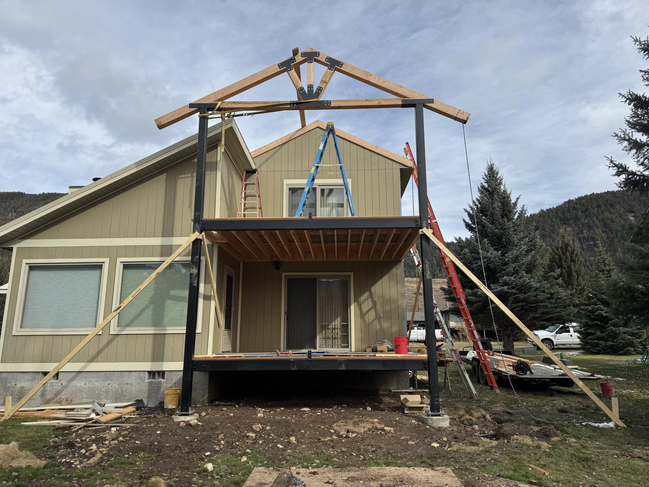 House under construction with a new deck and roof framework, ladders, and construction materials around.