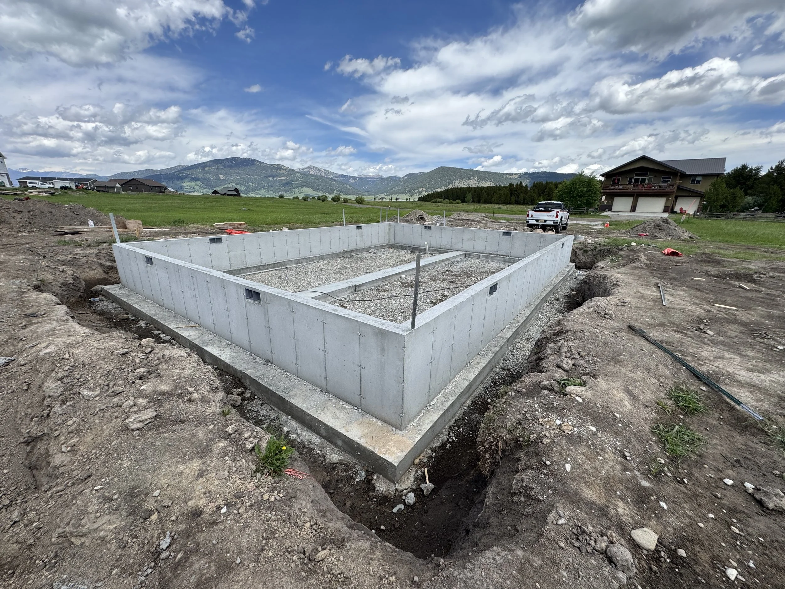 Concrete foundation for a building under construction in a rural area with mountains and houses in the background.