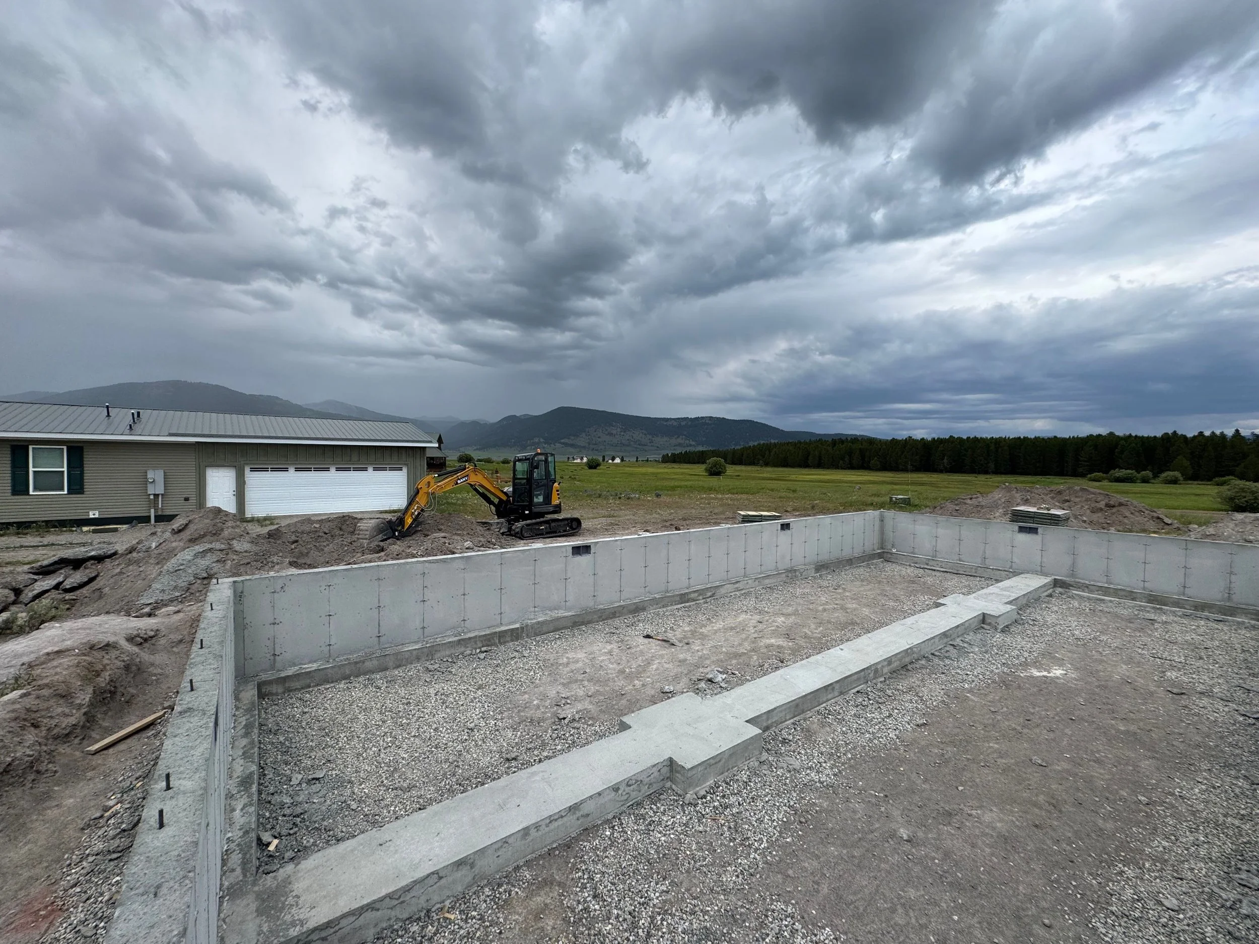 Construction site with concrete foundation, a small excavator, and a house in the background under a cloudy sky.