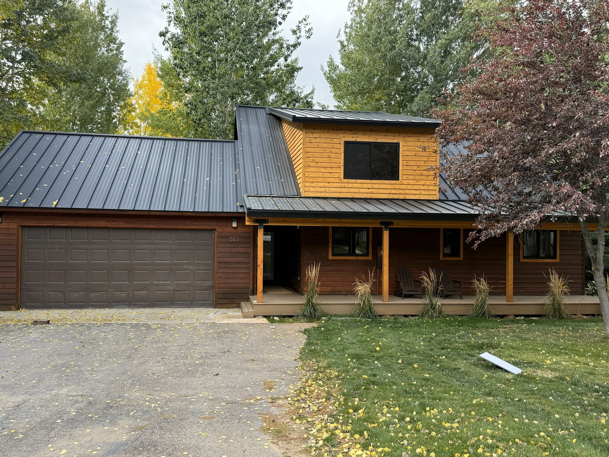 Front view of a modern house with a dark metal roof, wooden siding, and a small covered porch, surrounded by trees with fall foliage.