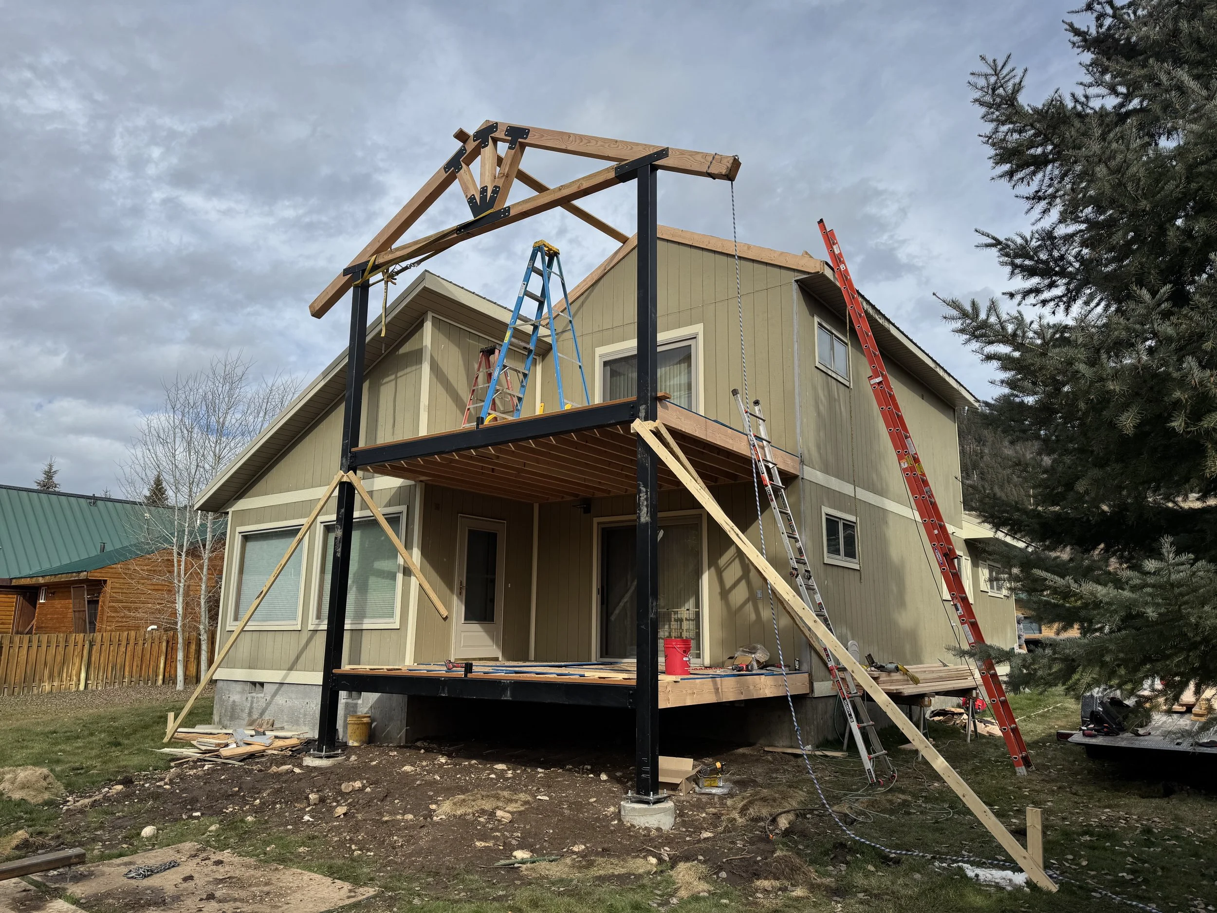 Construction of a two-story house, showing the framing and deck in progress with ladders and tools around.