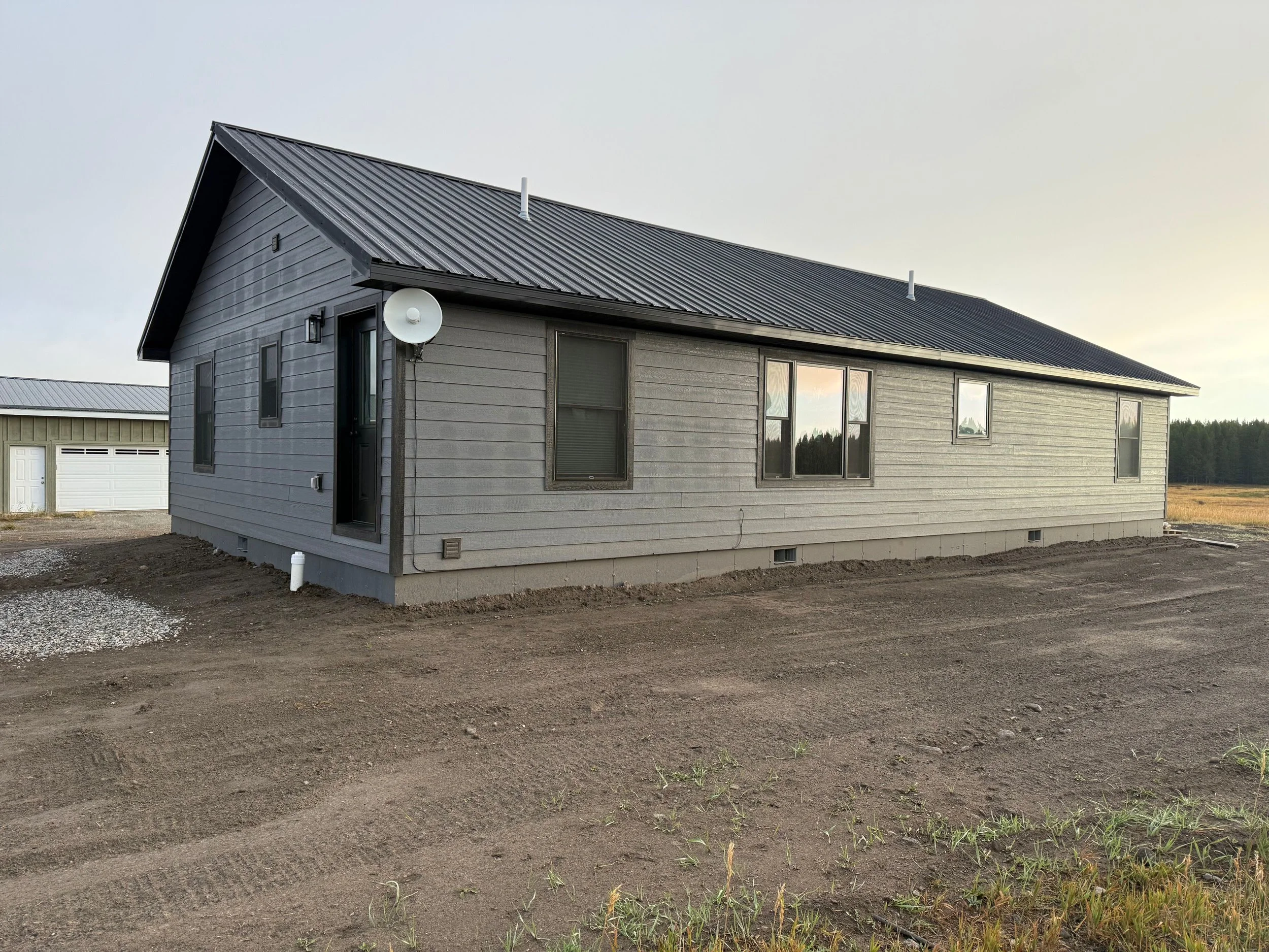 Exterior of a gray single-story house with a metal roof and multiple windows, situated on bare soil with a neighboring garage in the background.
