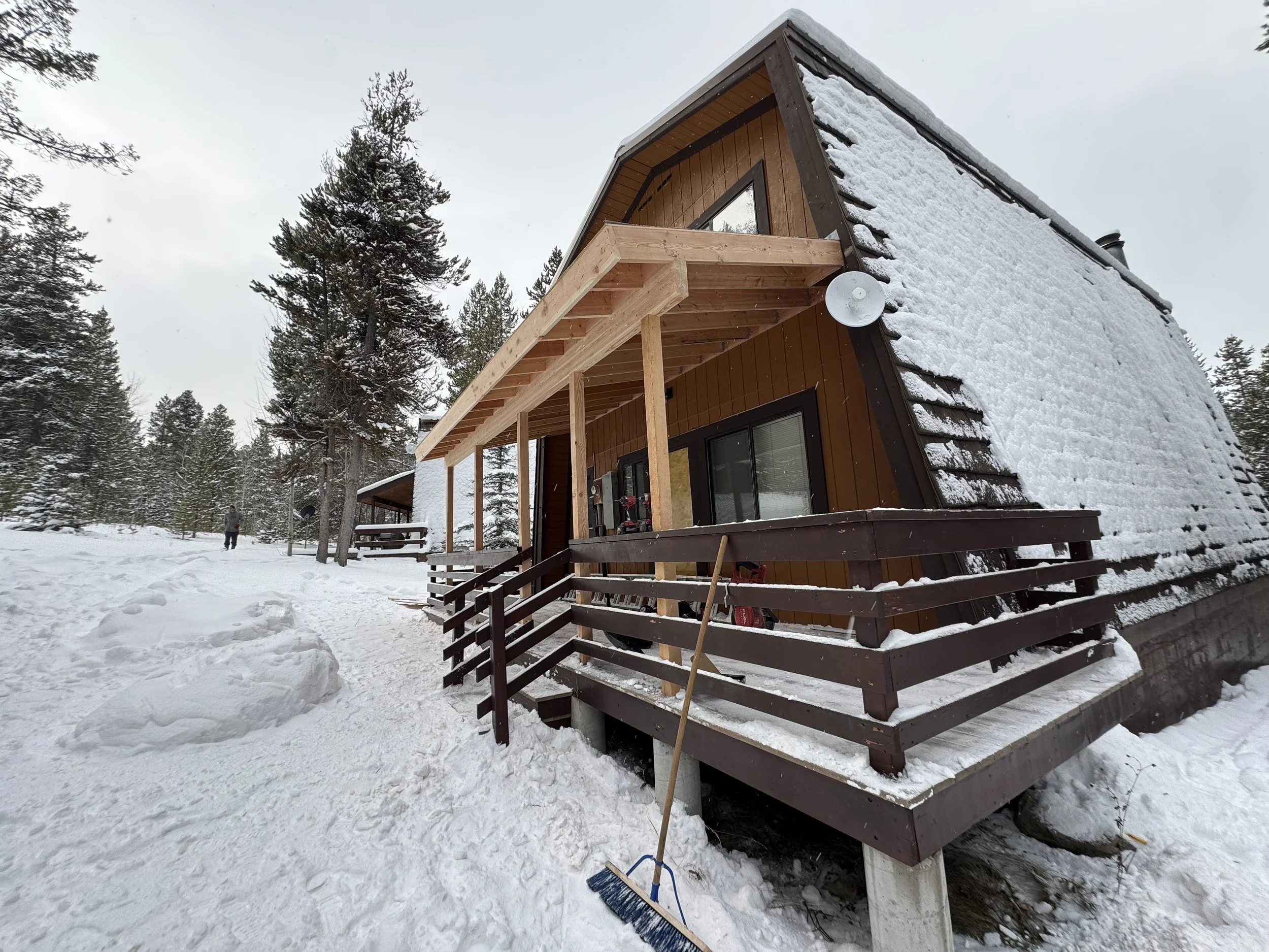 A snowy cabin with a large sloped roof, wooden exterior, and a front porch with railings. The hatch is covered in snow and surrounded by snow-covered trees.