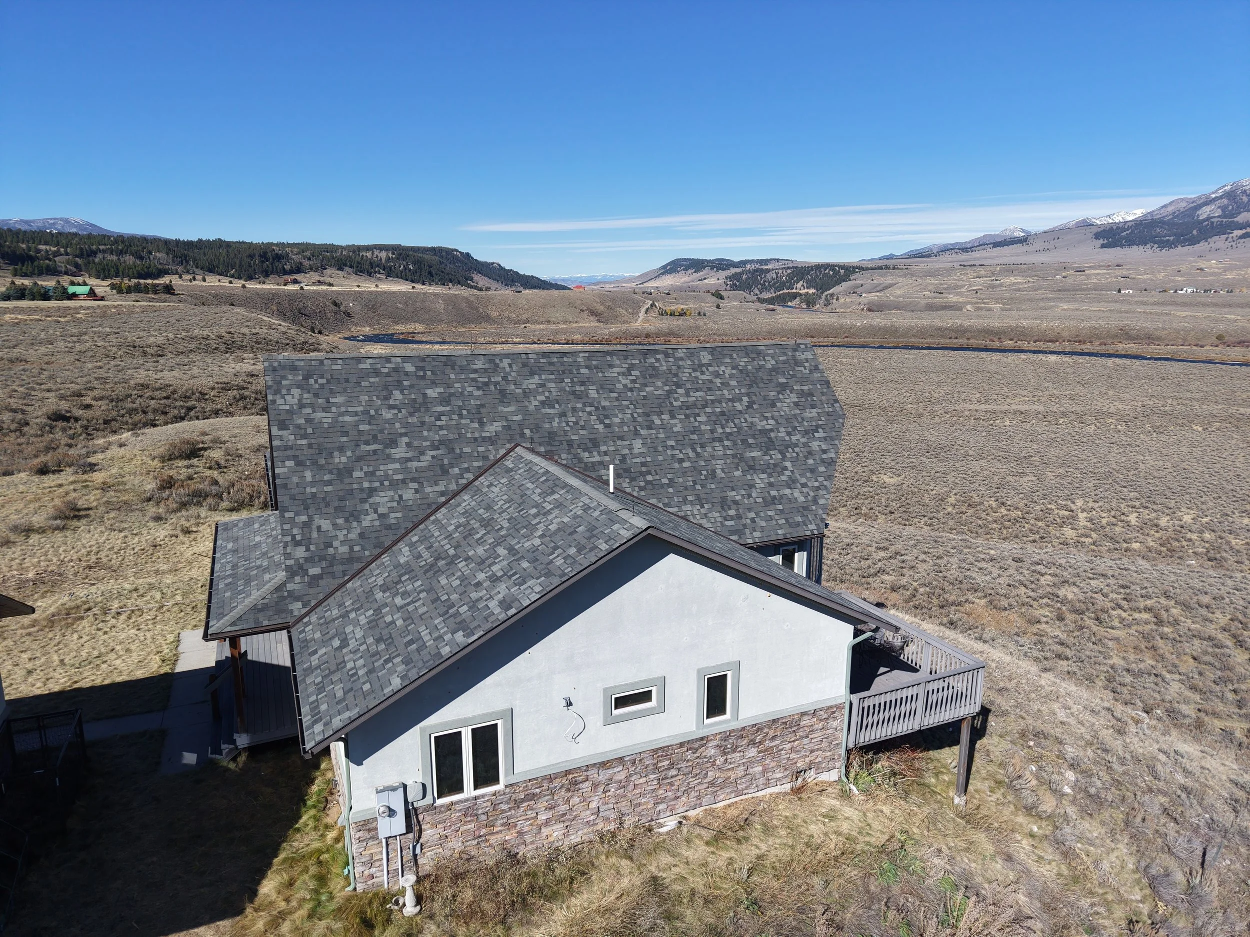 A house with gray shingled roof and white walls, situated on a dry, grassy landscape with mountains in the background under a clear blue sky.