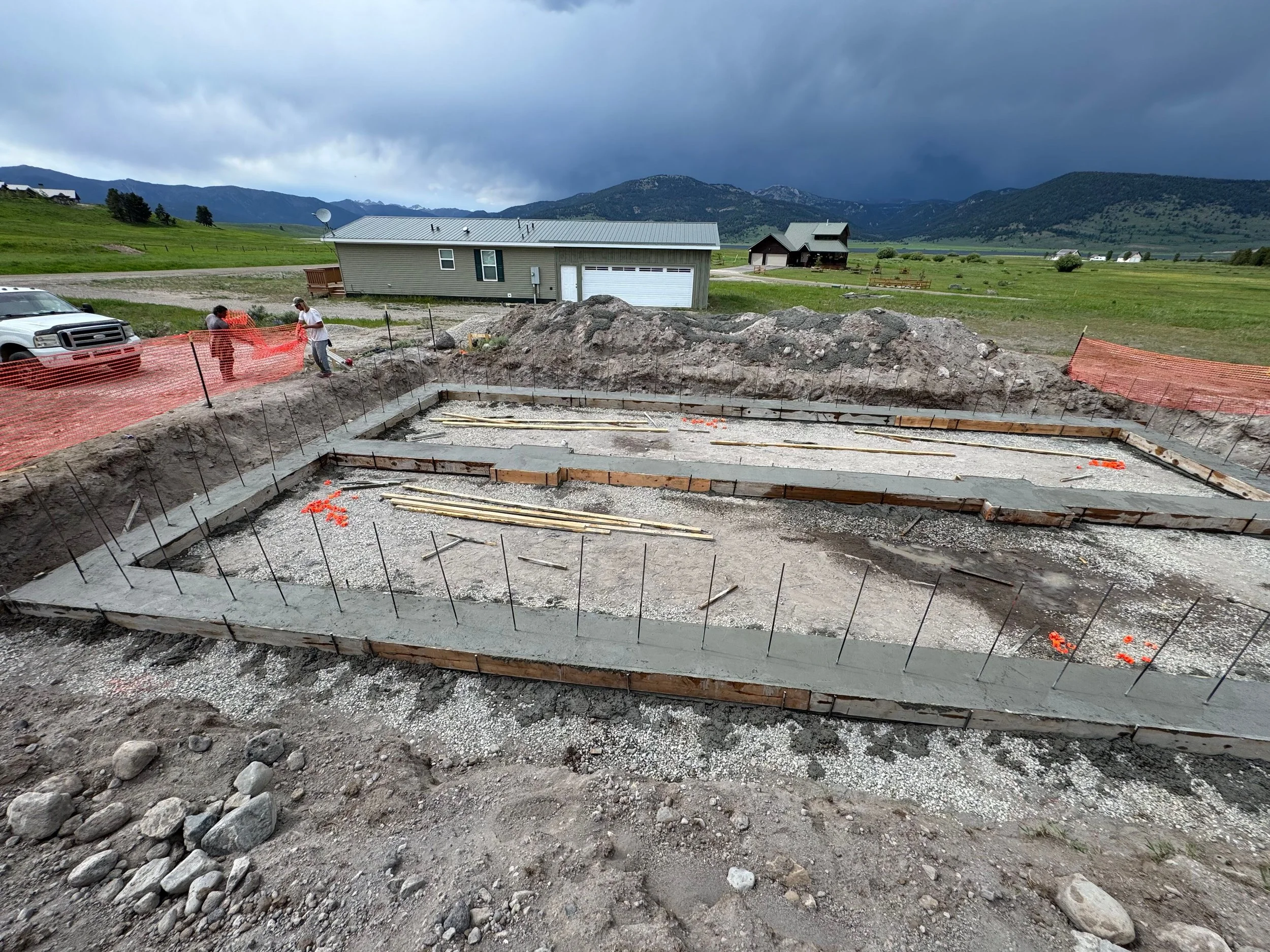 Construction site with concrete foundation being laid, orange safety fencing, workers, trucks, and rural landscape with green fields, houses, and mountains under dark cloudy sky.