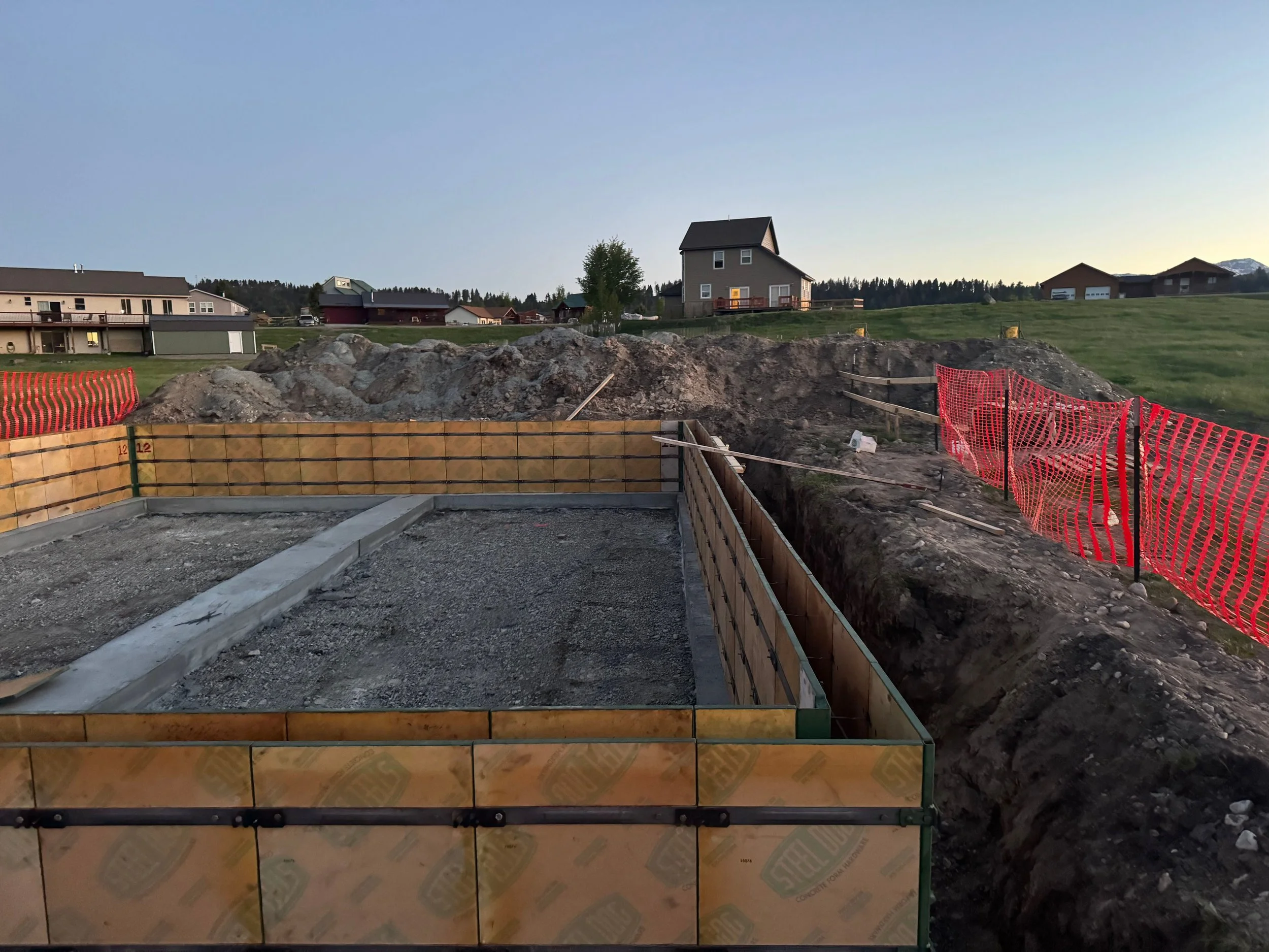 Construction site with a concrete foundation, orange safety fencing, and dirt piles in a residential area during dusk.