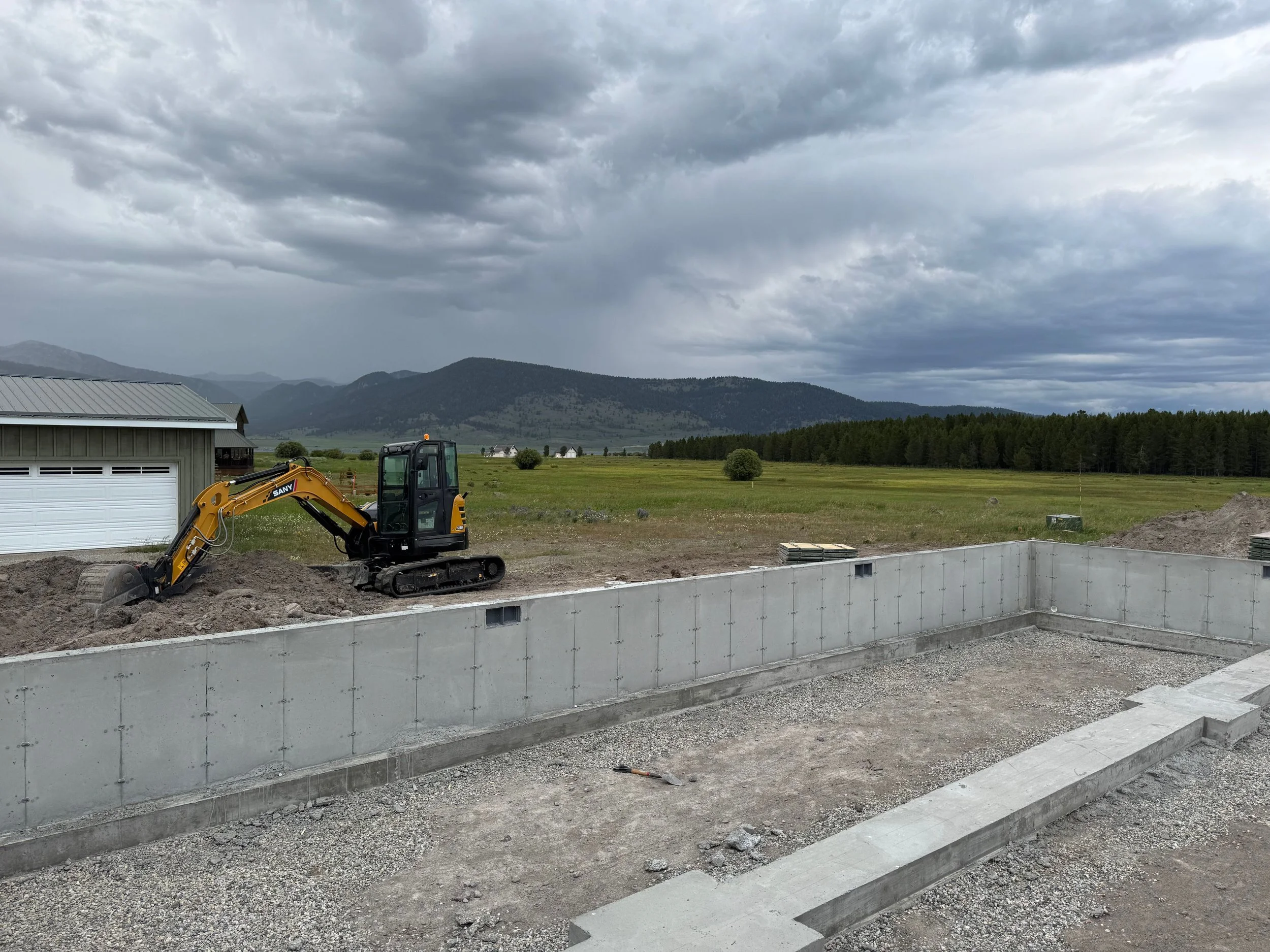 Construction site with a small excavator digging soil near a concrete foundation, open field with trees and mountains in the background under a cloudy sky.