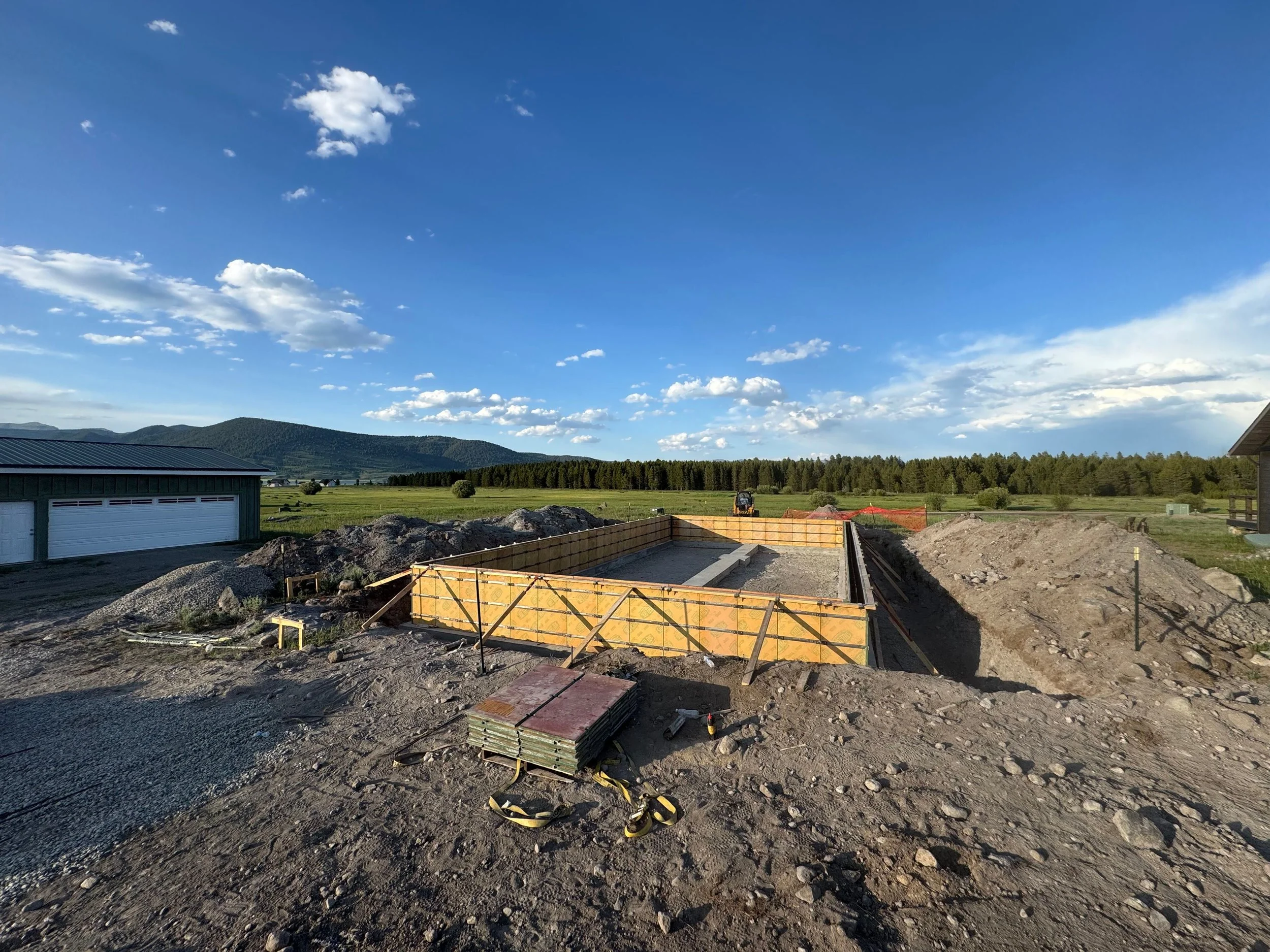 Construction site with wooden formwork set for a concrete foundation, piles of dirt and gravel around, in a rural area with green fields, trees, and mountains under a blue sky with clouds.