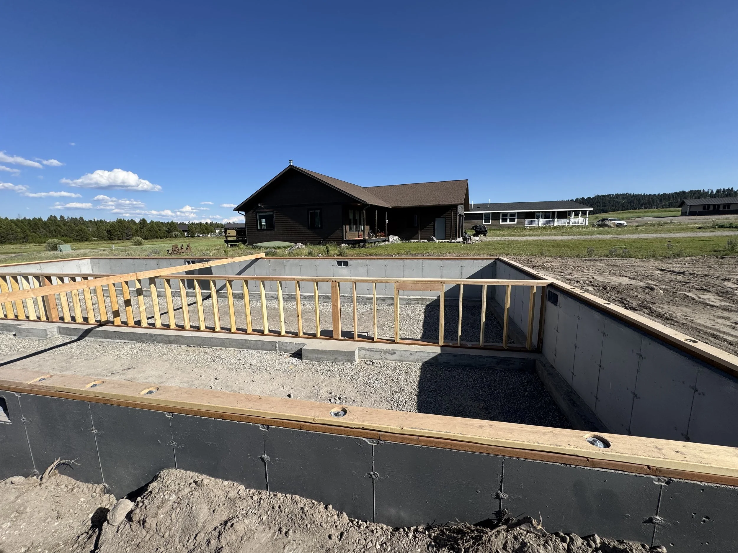 Construction site with building foundation and wooden framing in progress, set in a rural area with houses and open fields under a clear blue sky.