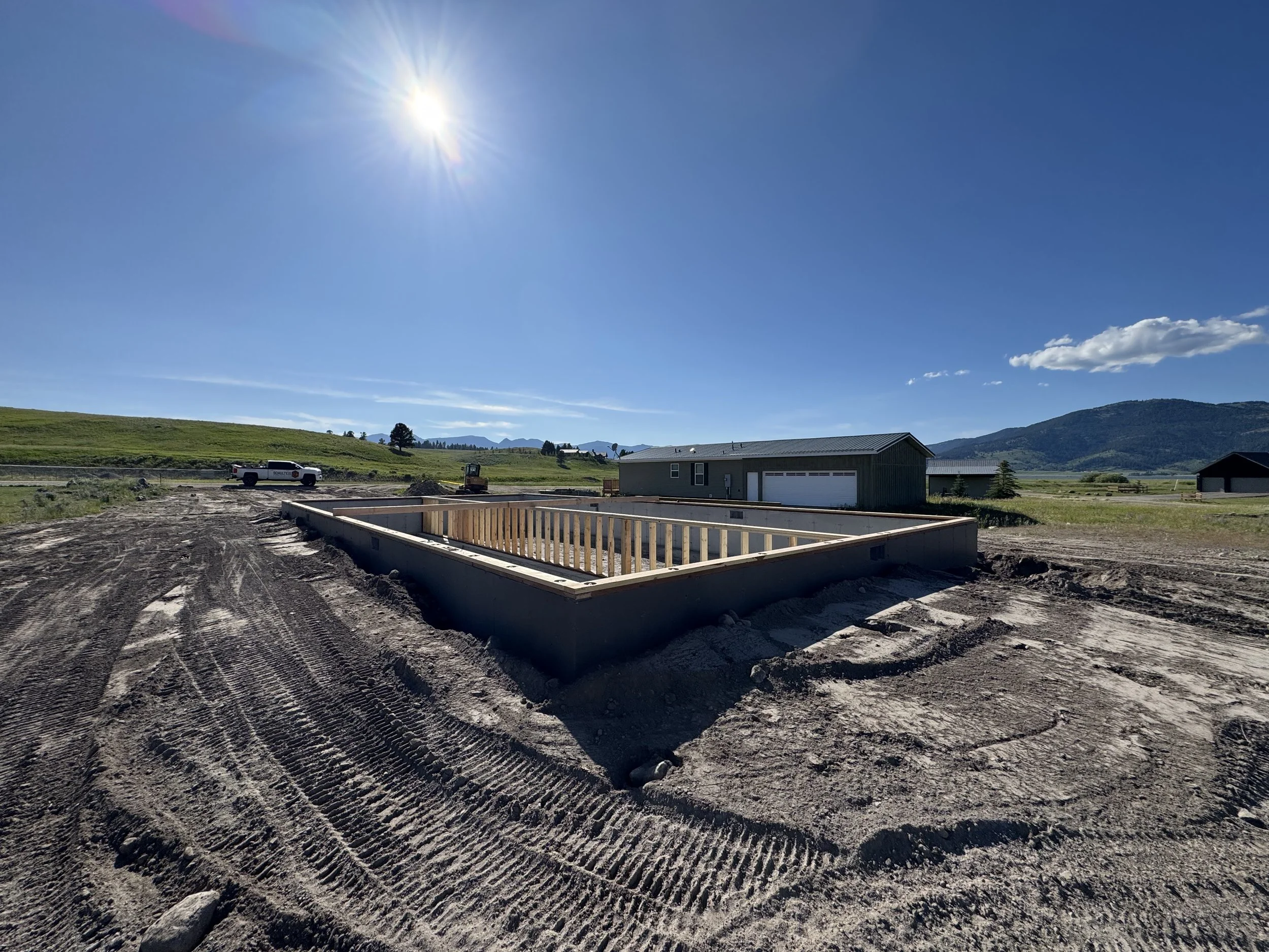 Construction site with a foundation outline and wooden frame, surrounded by dirt with tire tracks, under a clear blue sky with the sun shining, in a rural area with hills in the background.