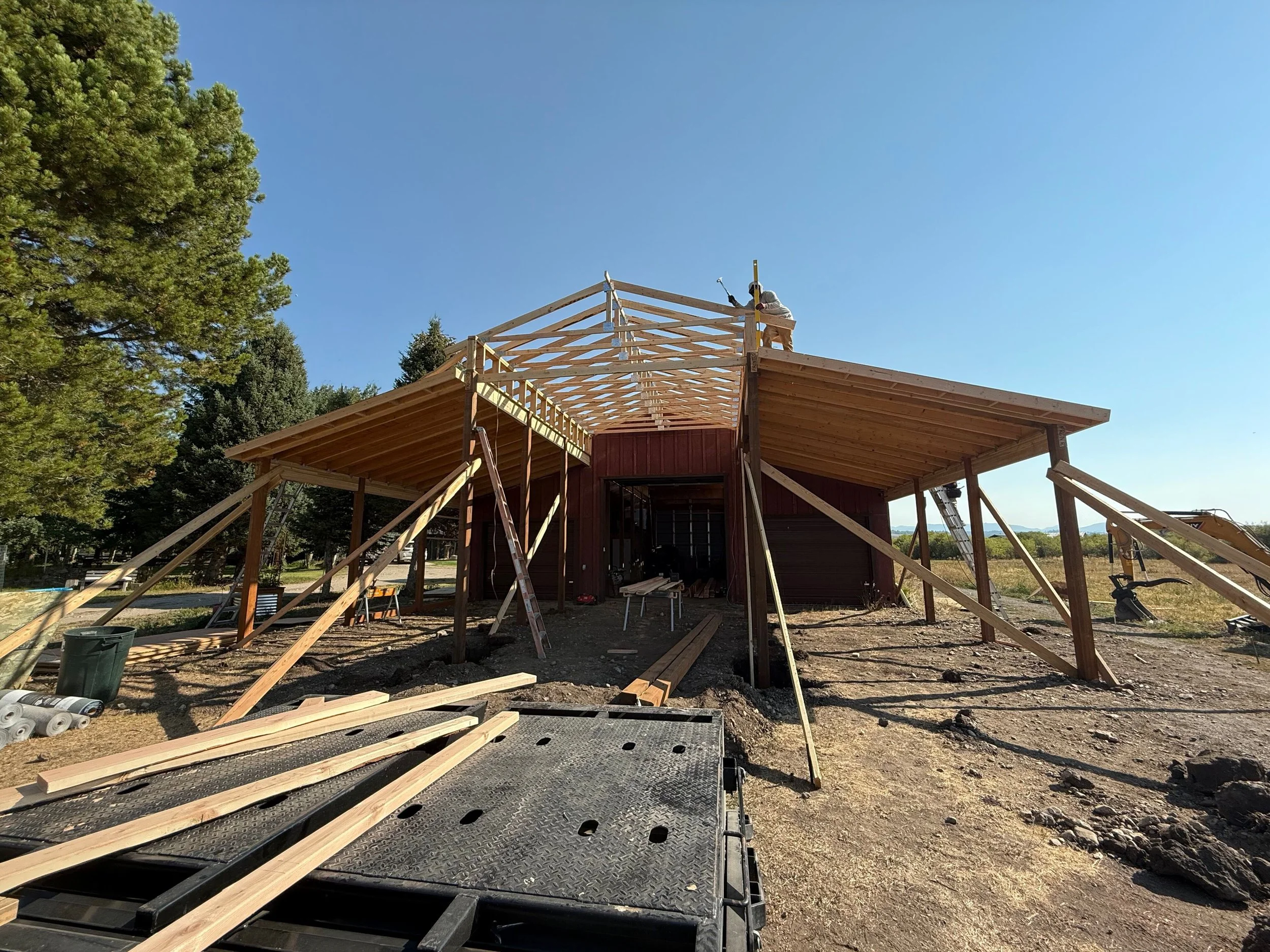Building under construction with wooden framework, scaffolding, and construction workers on the roof, surrounded by construction materials, in a rural area with trees and open sky.