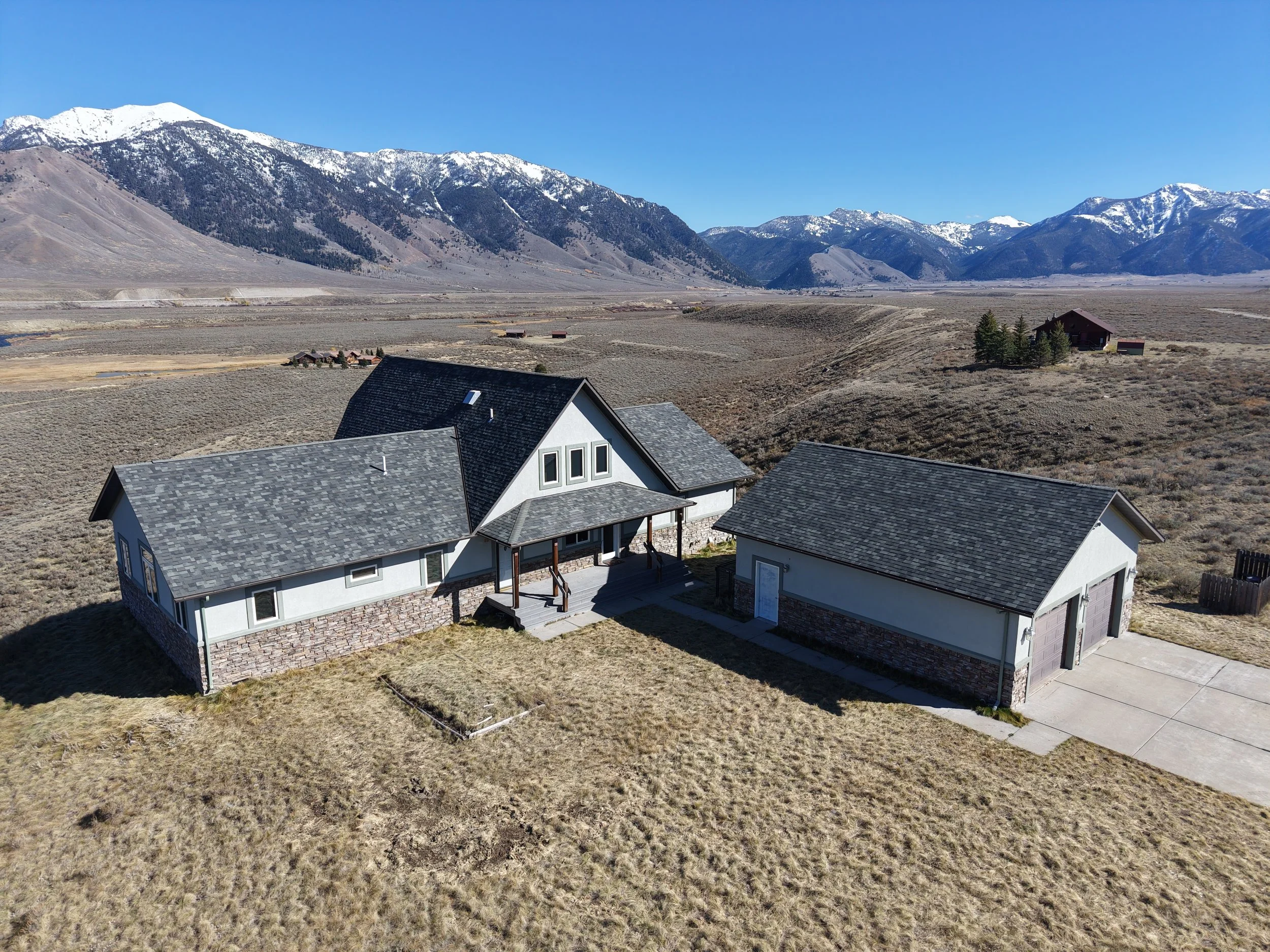 A house with a front porch and a detached garage in a rural, mountainous landscape with snow-capped mountains in the background.
