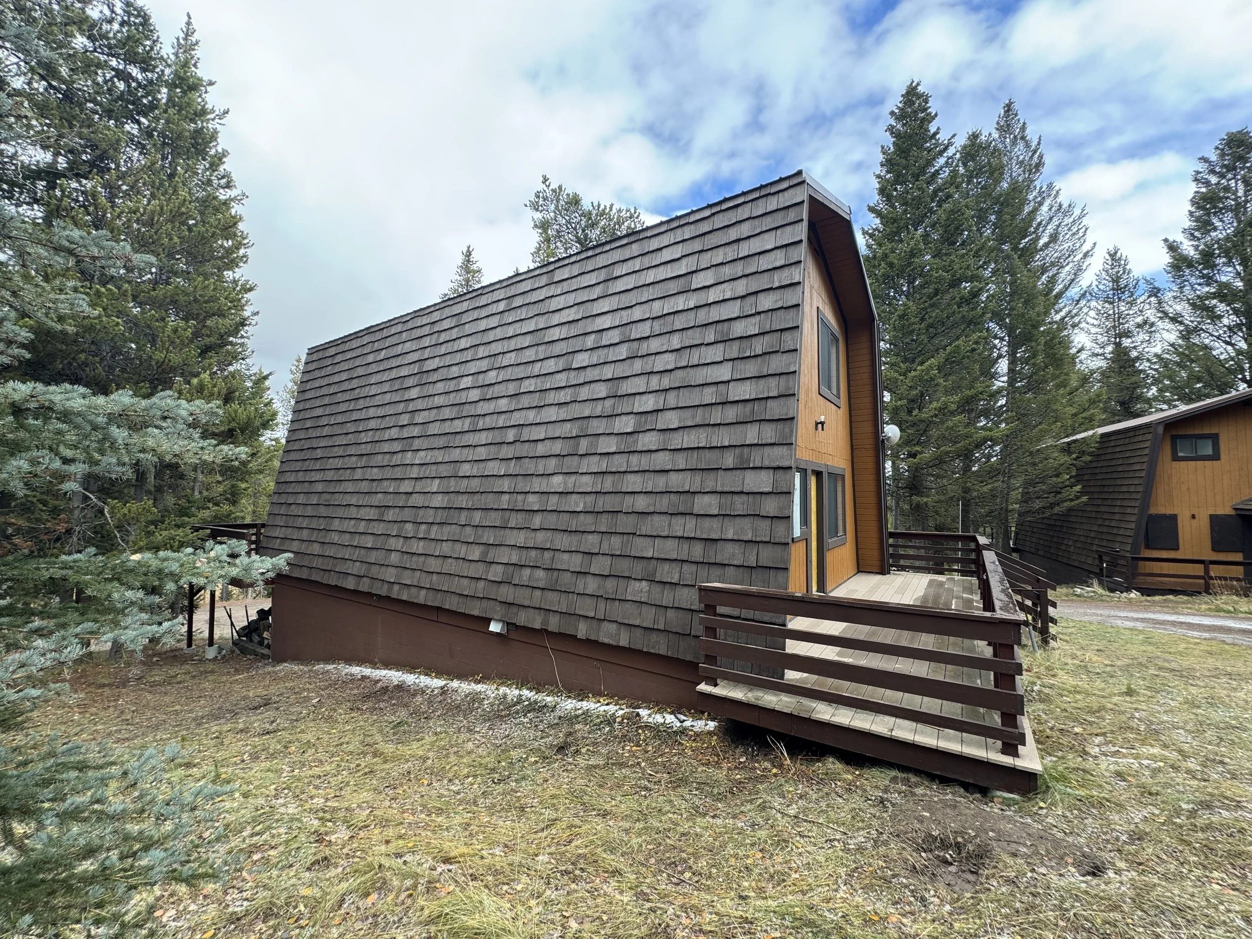 A brown A-frame cabin with a small wooden balcony, surrounded by pine trees and a partly cloudy sky.