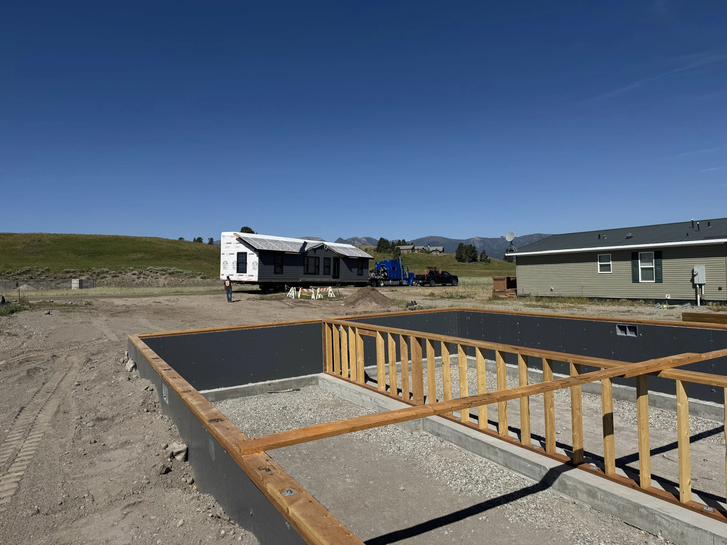 Foundation and framing for a house under construction with a partially built house on a trailer and a neighboring house in a rural area with mountains in the background.