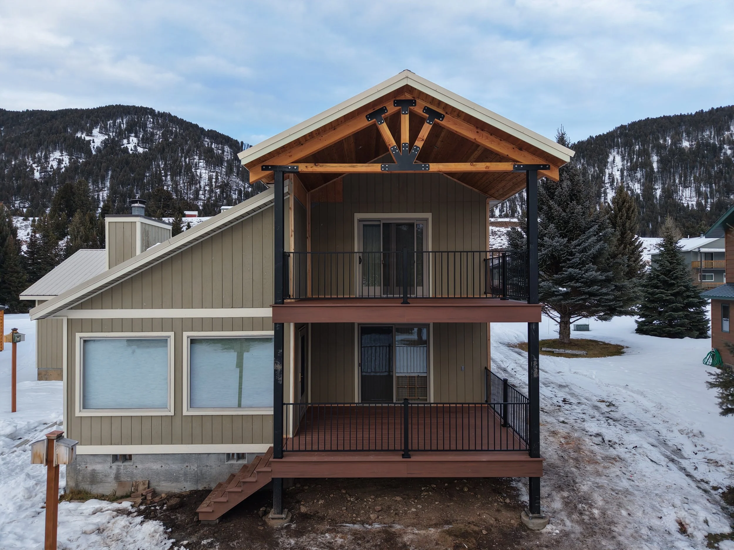 Two-story house with a wooden balcony, surrounded by snow, trees, and mountains in the background.