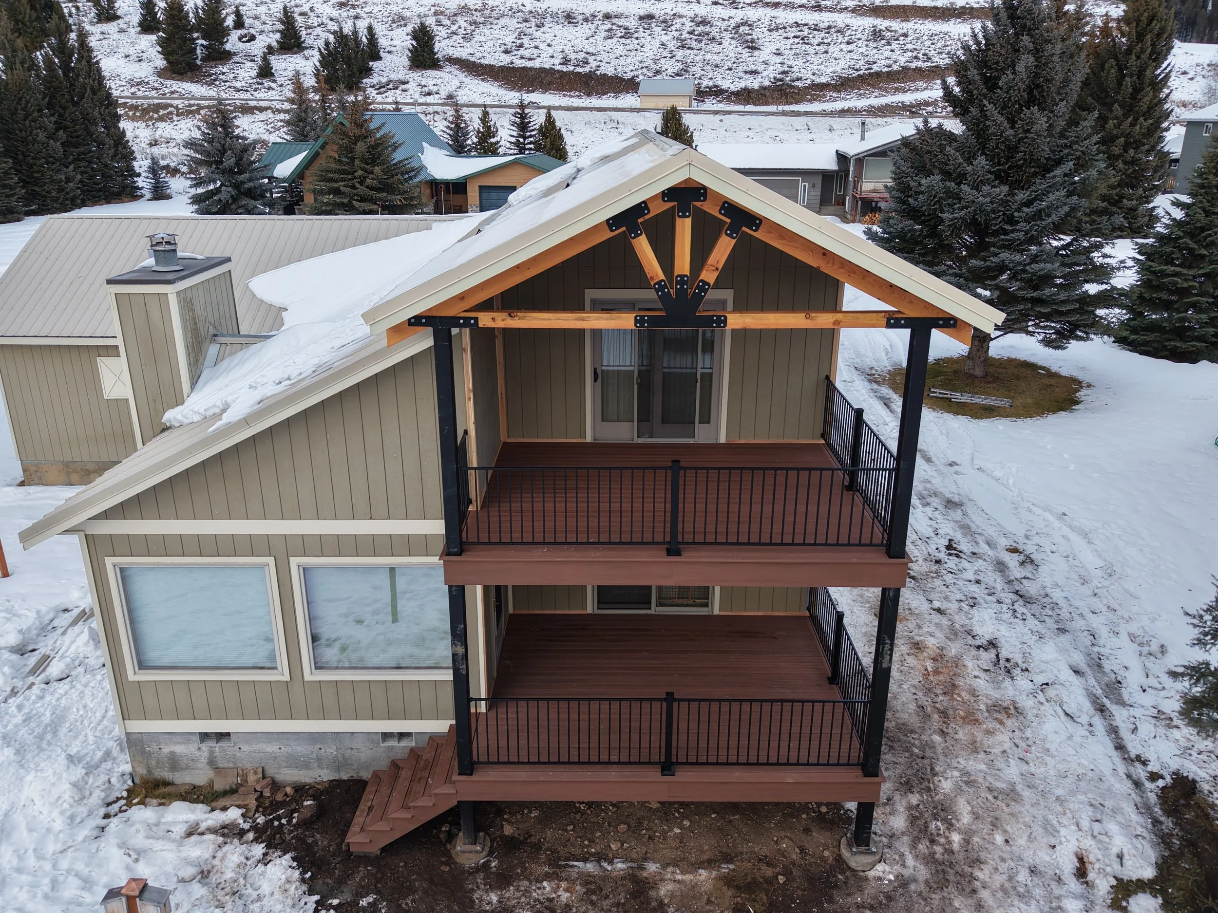 An aerial view of a two-story house with a covered front porch and a balcony on the upper level, with snow on the roof and ground, surrounded by trees and neighboring houses.